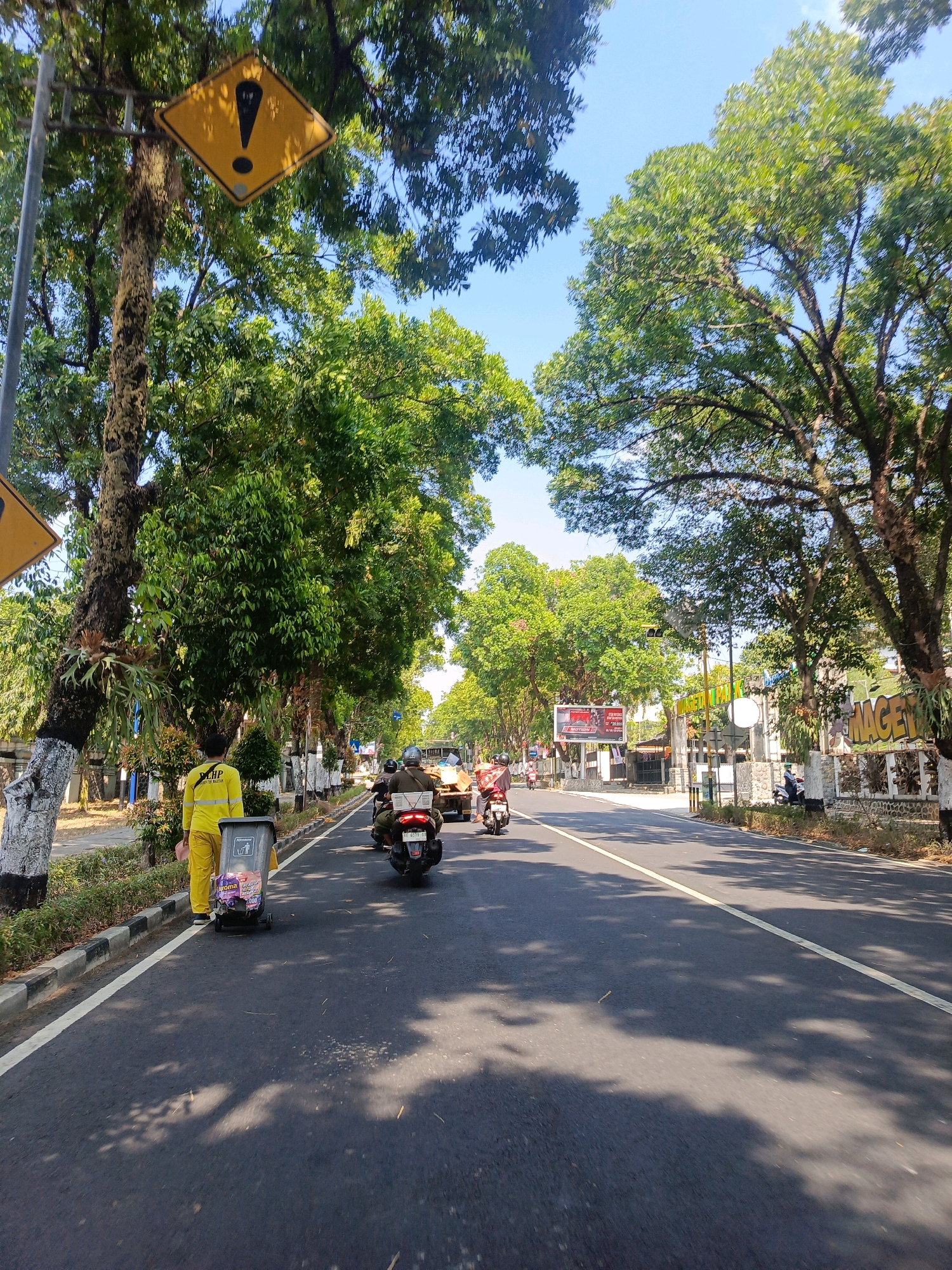 This is a medium-angle, eye-level shot of a tree-lined road in Tambran, Indonesia, under a clear blue sky. In the foreground, a person wearing a bright yellow uniform and pants, possibly a street cleaner, stands beside a grey trash bin on wheels. The trash bin contains several items, including what appears to be packaged snacks. The road is paved with asphalt, marked with a white line on the left side of the lane and a yellow line in the center.
Several motorcycles are visible on the road, with two in the immediate foreground and more further down. One motorcycle in the foreground has a rider wearing a helmet. In the background, there are more trees, buildings, and signs. One sign on the right side reads "MAGETAN" in large, yellow, three-dimensional letters. Another sign higher up on a pole, depicted in a close-up crop, is a yellow diamond shape with a black exclamation mark inside, indicating a warning. The trees lining the road have dense green foliage, and some have their trunks painted with white and black stripes. The overall impression is a sunny day on a moderately busy street in an Indonesian city.