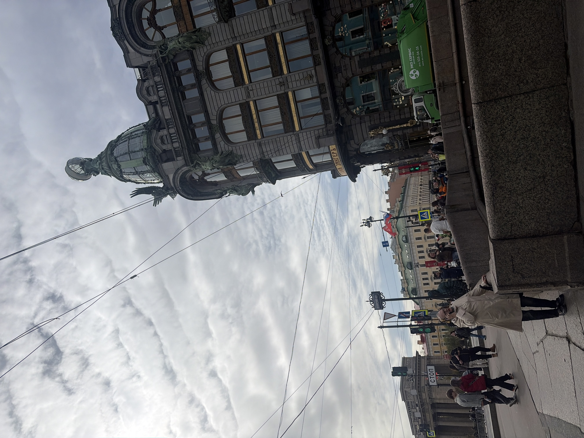 Here's a detailed description of the image:
**Scene Overview:**
The image captures a street scene in Saint Petersburg, Russia, specifically featuring the notable Singer House (Dom Knigi) building. The perspective is a low-angle, street-level view, partially tilted.
**Key Subjects and Objects:**
* **Singer House (Dom Knigi):** The prominent building is the focal point, showcasing its architectural features, including the glass dome topped with a globe, ornate facade, and sculptures.
* **Street Scene:** The image captures a bustling street with pedestrians, traffic lights, street signs (including "STOP" in Russian), and power lines.
* **People:** Pedestrians are visible on the sidewalk, including a person in a light-colored coat leaning against a wall. Other pedestrians are walking in various directions.
* **Vehicle:** A green truck is visible, possibly a service or delivery vehicle.
* **Russian Flags:** A few Russian flags are seen waving in the background.
* **Signage:** Street signs with names are visible.
**Environment:**
* **Sky:** The sky is overcast with cloudy weather conditions.
* **Architecture:** Other buildings are visible in the background, representing the architecture typical of Saint Petersburg.
* **Pavement:** The sidewalk is paved with stone.
**Activities and Interactions:**
* People are walking along the sidewalk.
* A person is leaning against a wall.
* A vehicle is parked or moving along the street.
* The presence of flags indicates a public space.