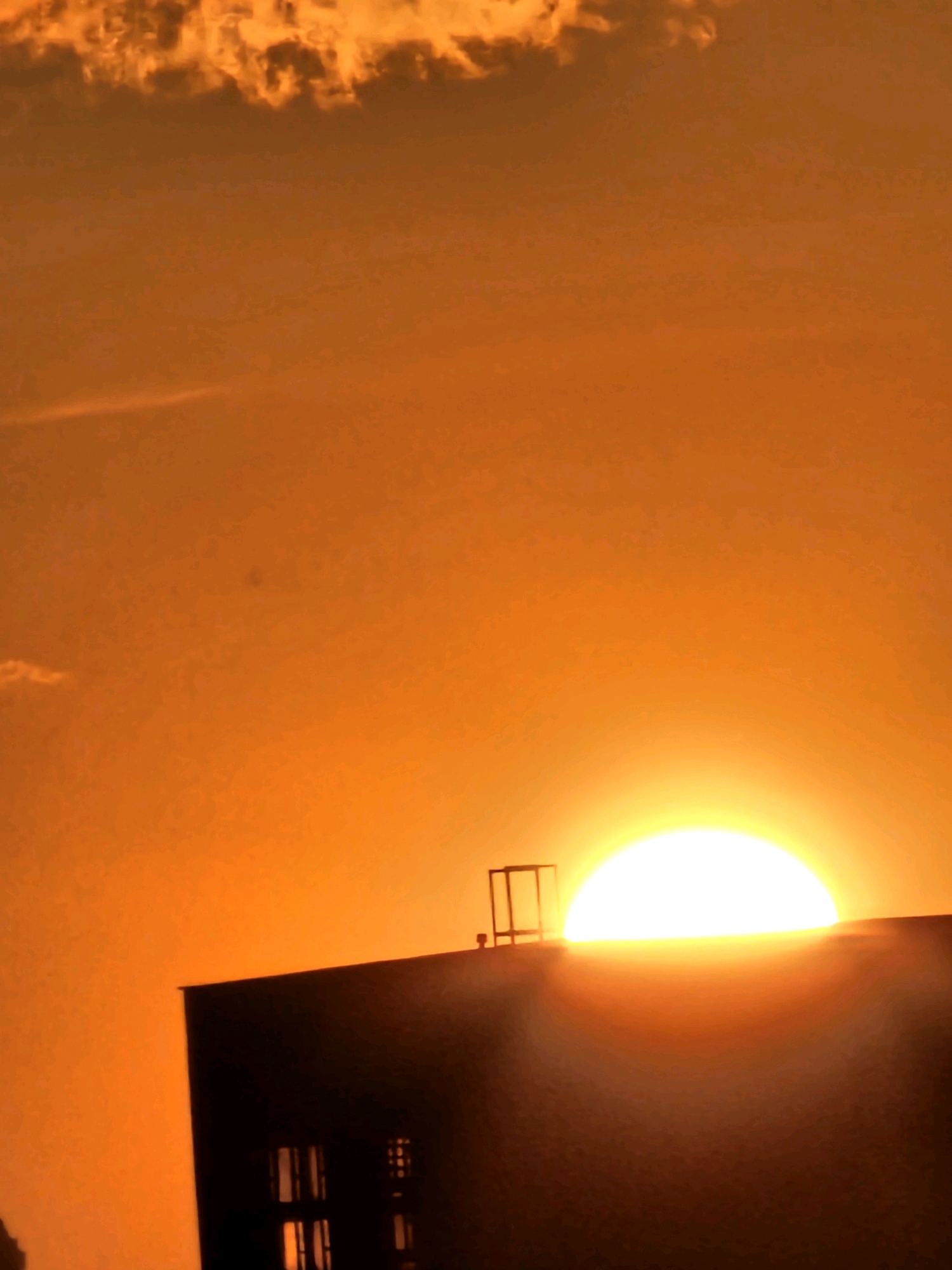 The image captures a sunset over the city of Khimki, Russia. The sky is a vibrant orange, with some clouds visible at the top. The sun, a bright, almost white orb, is partially obscured by the roof of a building, casting a warm glow. The building is dark, silhouetted against the sunset, with windows visible. A structure, possibly a ventilation shaft or a decorative element, is on the roof, also silhouetted. The scene is peaceful, with no people visible. The overall impression is one of a calm and serene evening in Khimki.