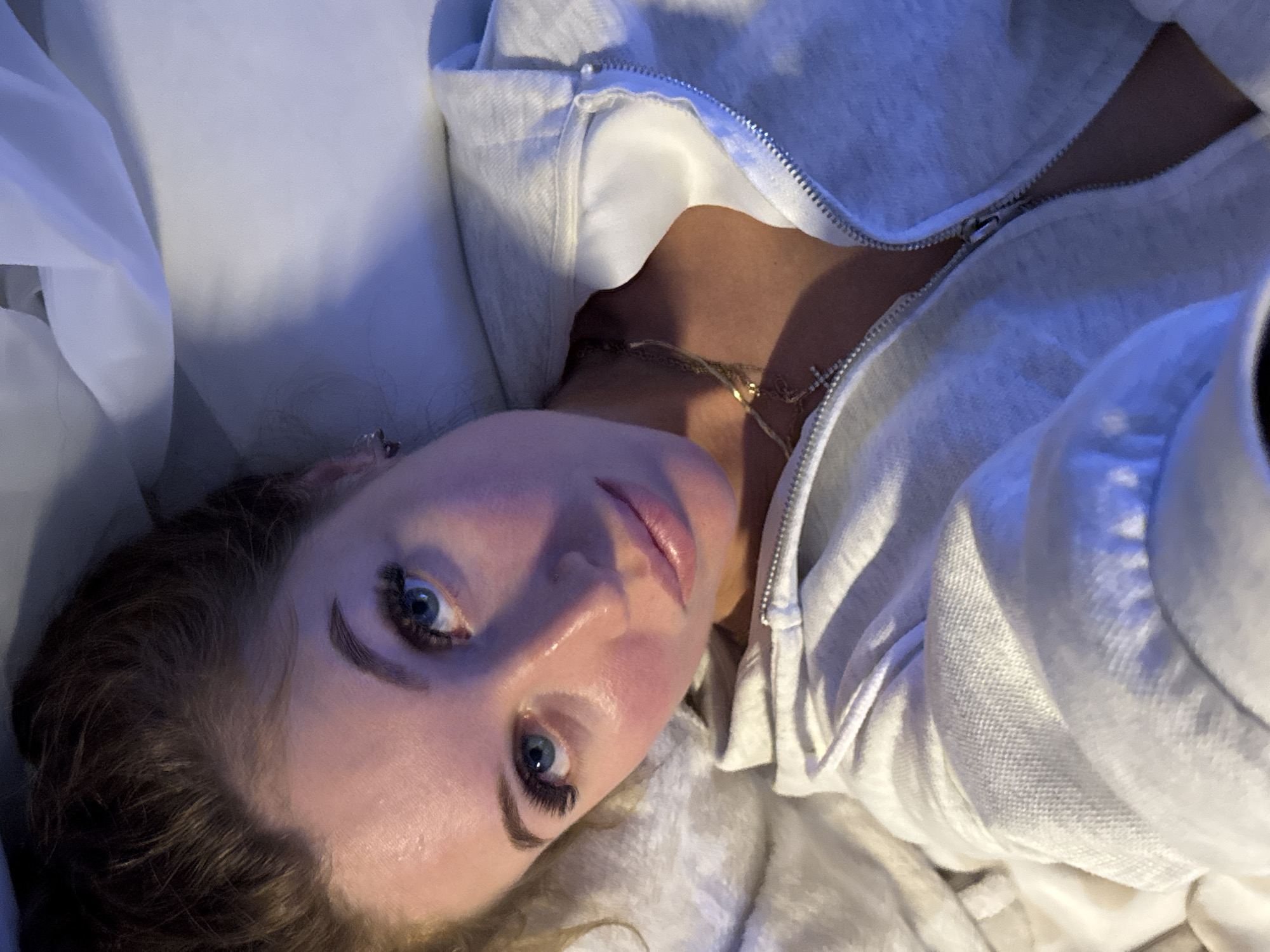 The image is a close-up, rotated selfie of a young woman lying down indoors. Her head is positioned towards the bottom left of the frame, with her body extending diagonally upwards.
The woman has fair skin, striking blue eyes with long, dark eyelashes, and well-defined eyebrows. Her lips are full and appear glossy. Her dark brown, wavy hair frames her face and shoulders. She is wearing a light-colored, possibly light grey or white, zip-up hoodie. Around her neck, a delicate, layered gold-colored necklace is visible, featuring a small cross pendant on one of its chains. She appears to be resting on soft, white or light-colored bedding or fabric.
The lighting is soft and has a distinct cool, blueish tint, indicative of artificial light and suggesting the photograph was taken indoors, likely in the evening or at night. Her expression is calm and direct as she gazes into the camera, implying a relaxed or posed selfie. There are no specific visual cues within the image itself to confirm the specified location of Chaikovsky, Russia. No text is visible in the photograph.