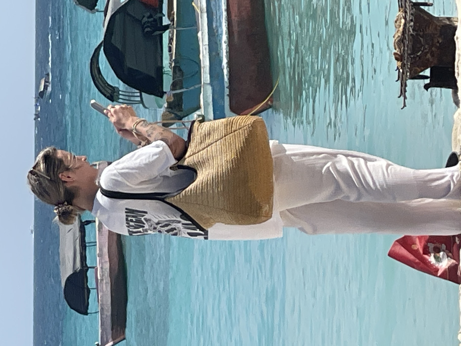 The image depicts a coastal scene in a shallow, turquoise body of water, likely a beach or waterfront area in Zanzibar, Tanzania. The central subject is an individual, appearing female, positioned with their back slightly turned towards the camera, looking down at a silver smartphone held with both hands.
The person has blonde or light brown hair tied back in a bun and is wearing an oversized white t-shirt with a black and white graphic on the back depicting an animal or skeleton and the partial text "THE OCEAN." They wear white, loose-fitting trousers and dark flip-flops or sandals. A large, light brown woven straw tote bag with black straps hangs over their left shoulder. Several thin bracelets are visible on their right wrist, and sunglasses rest on top of their head. A tattoo is visible on the left arm.
The water behind the individual is clear and light blue, indicating shallow conditions. Several wooden boats with canopies are present in the water. To the left, a reddish-pink boat with a dark, arched canopy floats. To the right, another boat, partially obscured, shows blue and reddish-brown hull sections with a similar dark canopy. In the distant background, several more boats are visible on the horizon, one appearing to be a speedboat generating a wake.
On the sandy foreground, near the individual's feet, a crumpled red object, possibly a bag or fabric, is visible to the left. To the right, a makeshift metal grill or barbecue stand with what appears to be food, possibly grilled seafood, is positioned on the sand. The sky is clear and bright.