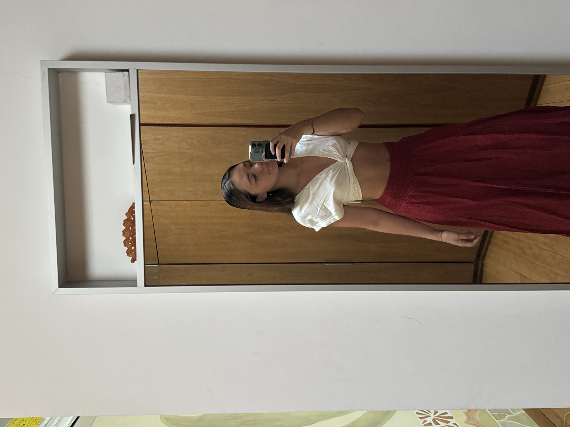 A woman is taking a mirror selfie inside a room. The woman is wearing a white, short-sleeved crop top and a red skirt. She holds a black mobile phone in her left hand. The mirror has a silver frame and is reflecting a wooden closet. The surrounding walls are white and decorated with floral patterns. The location is in San Bernardino Tlaxcalancingo, Mexico.