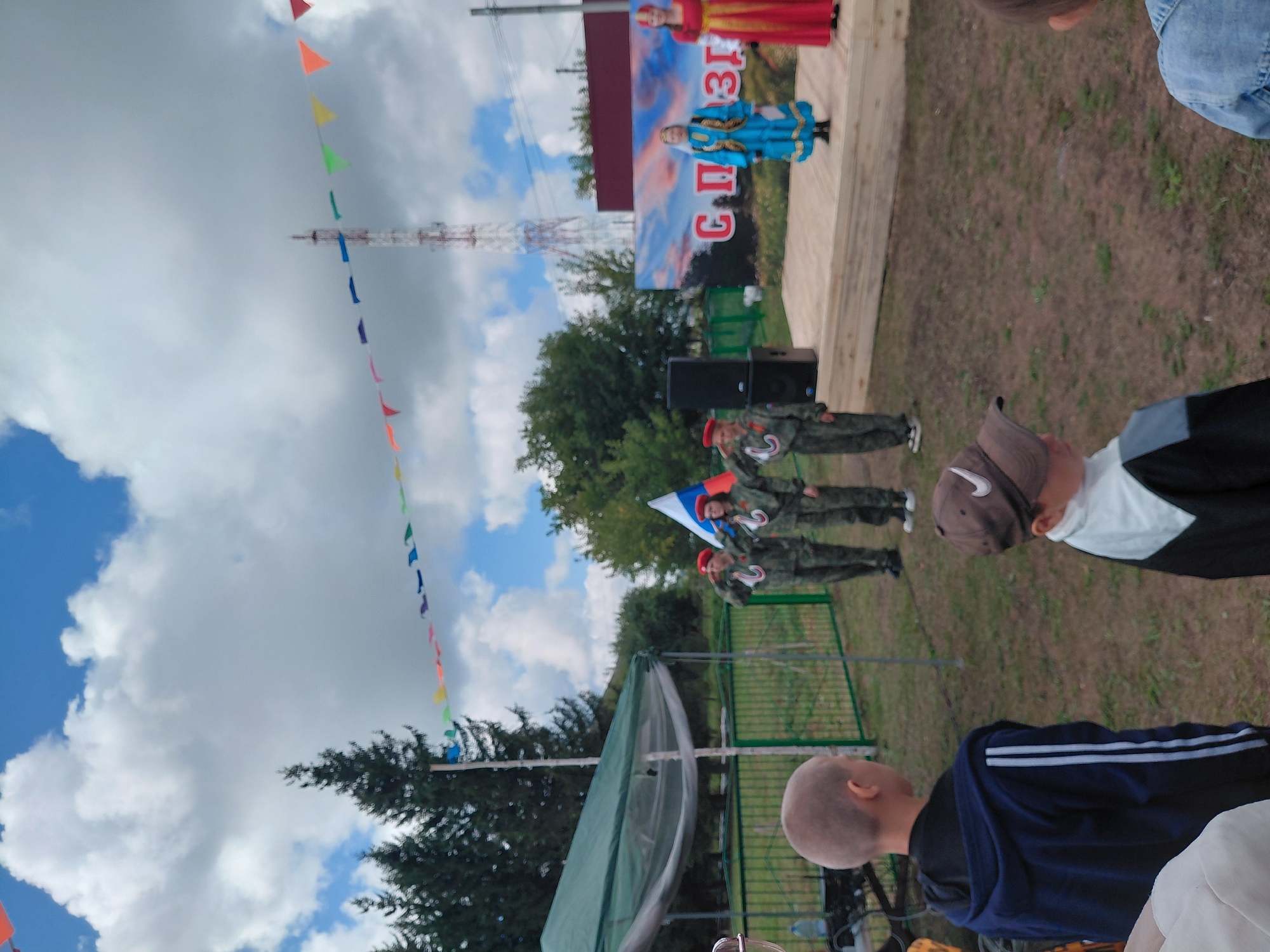 This is a medium-distance, eye-level, full shot of an outdoor event, likely in Novosyolovo, Russia. The scene features a group of people, including children, gathered on a grassy area in front of a wooden stage. Three children in military-style uniforms and red berets stand at attention on the stage, saluting a Russian flag. To their right, two women in traditional Russian attire, one in a blue dress and the other in a red dress, are also on the stage. A banner with Russian text and a sunset image is visible behind them. In the foreground, two young boys are facing away from the camera, observing the event. A green tent and a fence are to the left of the stage. The sky is partly cloudy, with a communication tower visible in the distance. The event appears to be a local celebration or ceremony.
