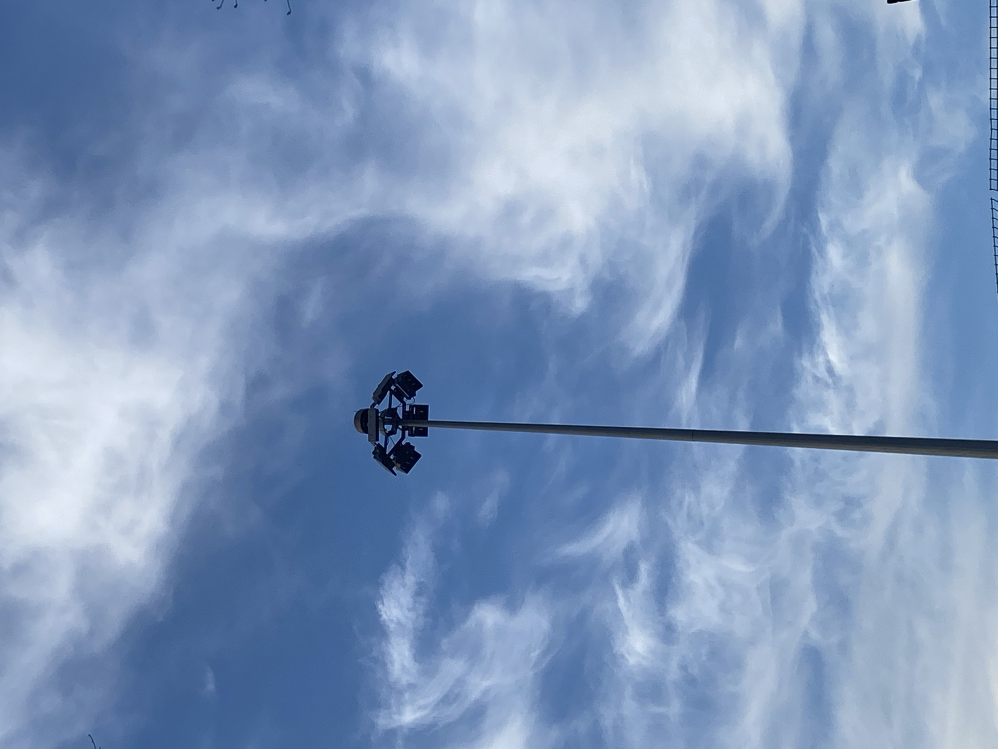 The image displays a single, tall, grey high-mast lighting pole centrally positioned against a blue sky with scattered white, wispy clouds. The pole supports a circular head assembly equipped with approximately eight to ten dark, rectangular floodlight fixtures, angled downwards. The sky exhibits varying shades of blue with distinct, elongated cloud formations. A dark, grid-like structure is partially visible along the bottom right edge of the frame, suggestive of a fence or netting. No human subjects, animals, or vehicles are present. The scene is illuminated by natural daylight. This high-mast lighting structure is located in Khimki, Russia.