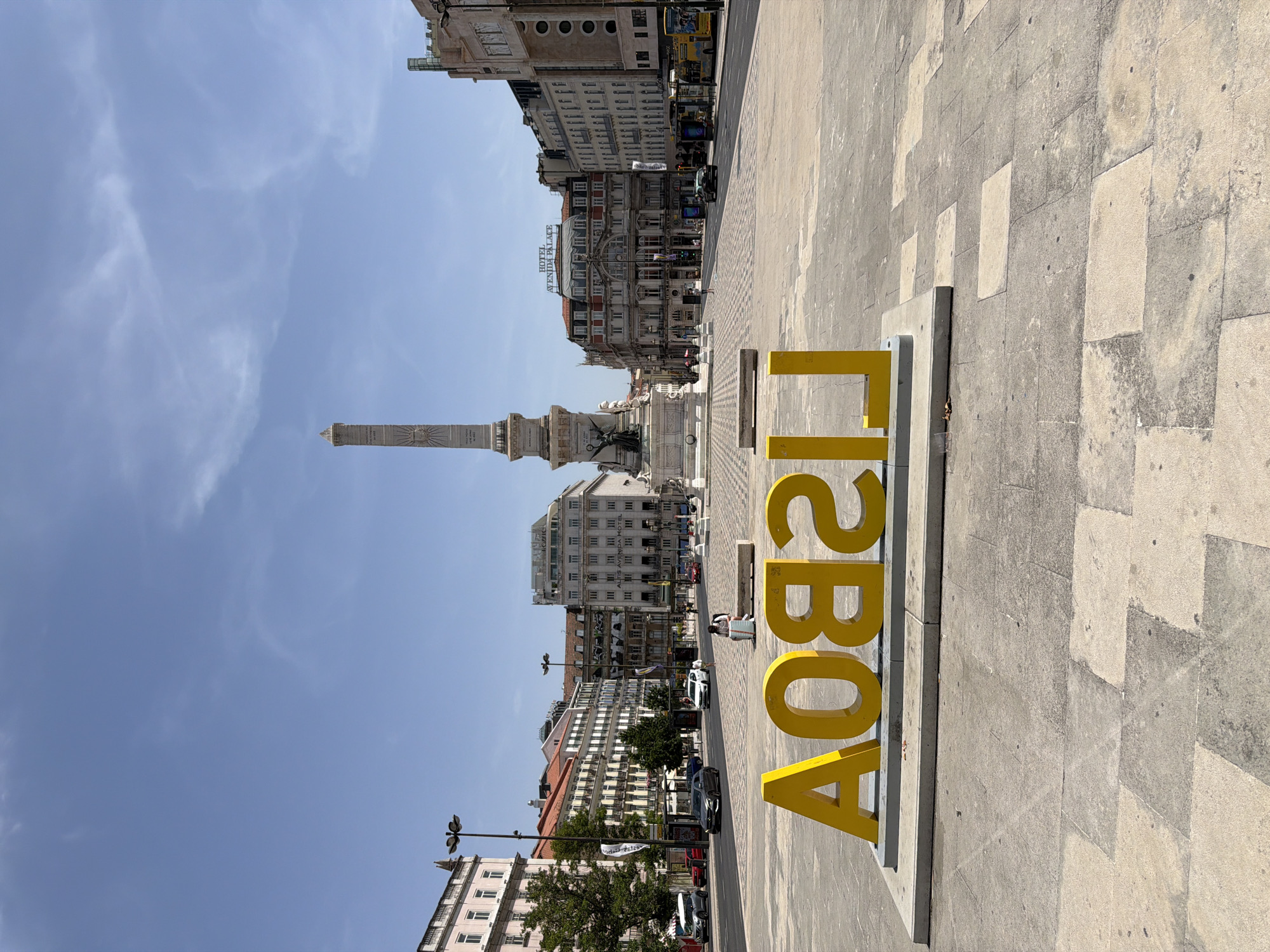 The image depicts a sunny daytime scene in Lisbon, Portugal, specifically at Rossio Square. In the foreground, large, bright yellow, three-dimensional letters spell out "LISBOA" (Lisbon) in reverse. These letters are positioned on a concrete platform. Behind the letters, the square opens up to a wide, paved plaza with a distinctive patterned stone surface. A white bench is visible behind the "LISBOA" sculpture.
Further into the background, a prominent obelisk, the Monument to King Pedro IV, stands at the center of the square. It is surrounded by ornate buildings, including the Hotel Avenida Palace, which is easily identifiable by its signage. The buildings exhibit classical architecture with multiple stories and arched windows.
The street running through the square is active with traffic, including several cars and yellow buses. Bus stops are visible on the sides of the street. A few pedestrians can be seen walking or sitting on benches. The sky is clear blue with some wispy white clouds. The overall impression is a vibrant and bustling urban square on a clear day.