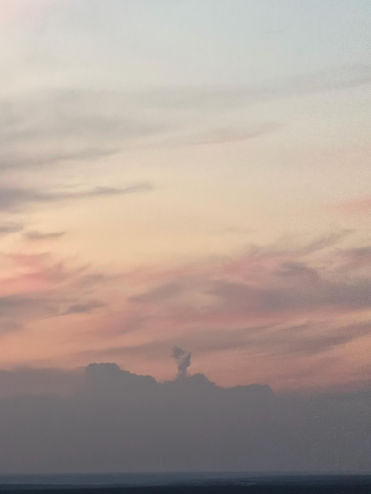 A wide shot captures a hazy sunset over Kyiv, Ukraine. The sky transitions from a pale blue at the top to soft pinks and oranges near the horizon, with wispy clouds scattered throughout. In the foreground, dark, silhouetted clouds form a jagged line against the colorful sky. The lower portion of the image shows a faint, dark landscape, possibly trees or buildings, with a thin line of darker blue indicating water or a distant horizon.