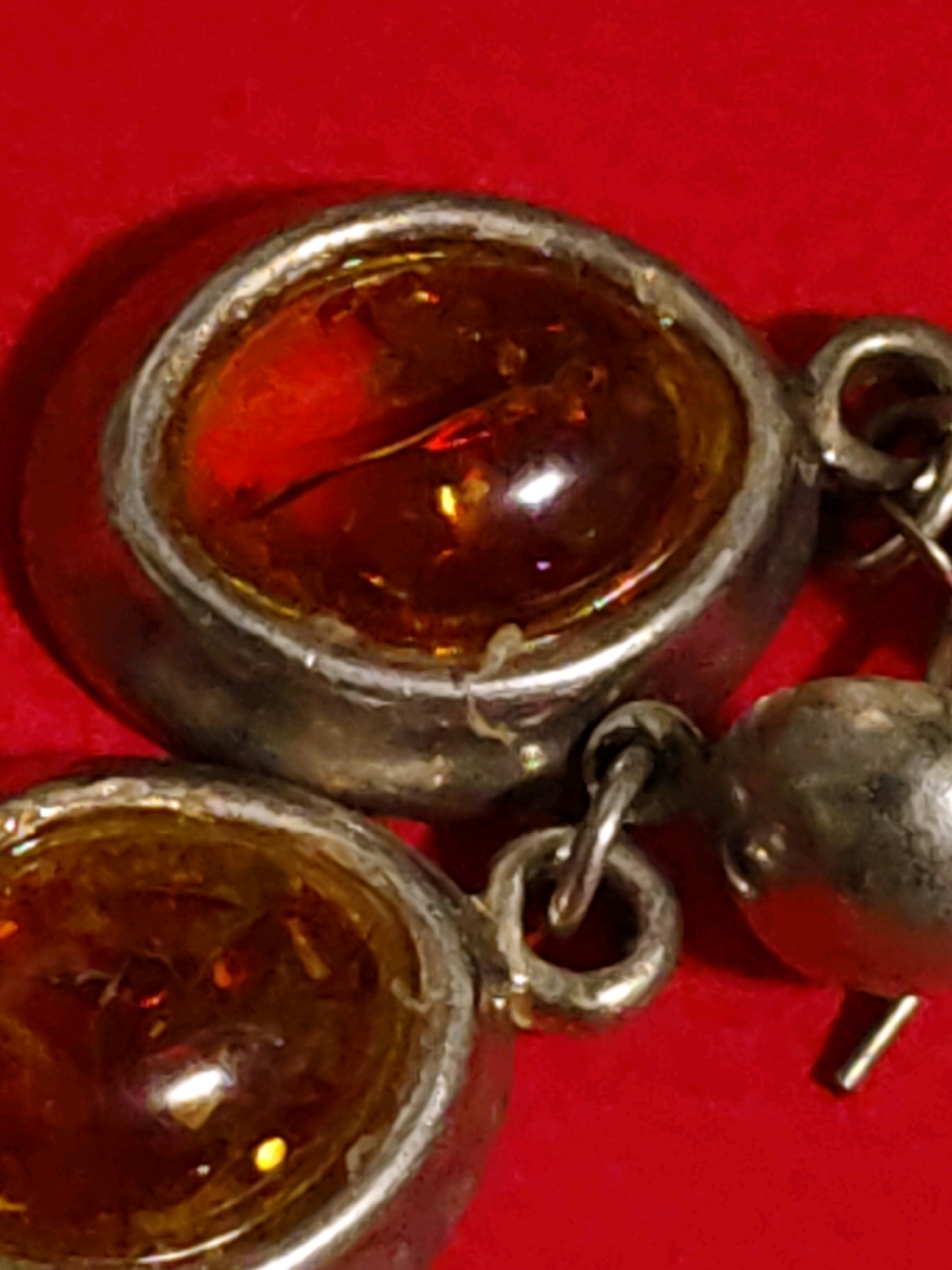 The image is a close-up, high-angle shot of jewelry, likely a bracelet or necklace, against a red, textured background. The jewelry features two circular amber stones set in a metallic, possibly silver, bezel. The amber stones are translucent with a deep orange-red hue, exhibiting some internal inclusions and reflections of light. The metalwork appears somewhat aged or antique, with visible texture and slight imperfections. Connecting the amber settings are small, metallic links and a charm that resembles a small bell or bead. The lighting creates strong shadows, particularly behind the main amber stone, and highlights the reflective surfaces of the amber and metal. There are no people or any discernible elements that would indicate a location in Rostov-on-Don, Russia.
