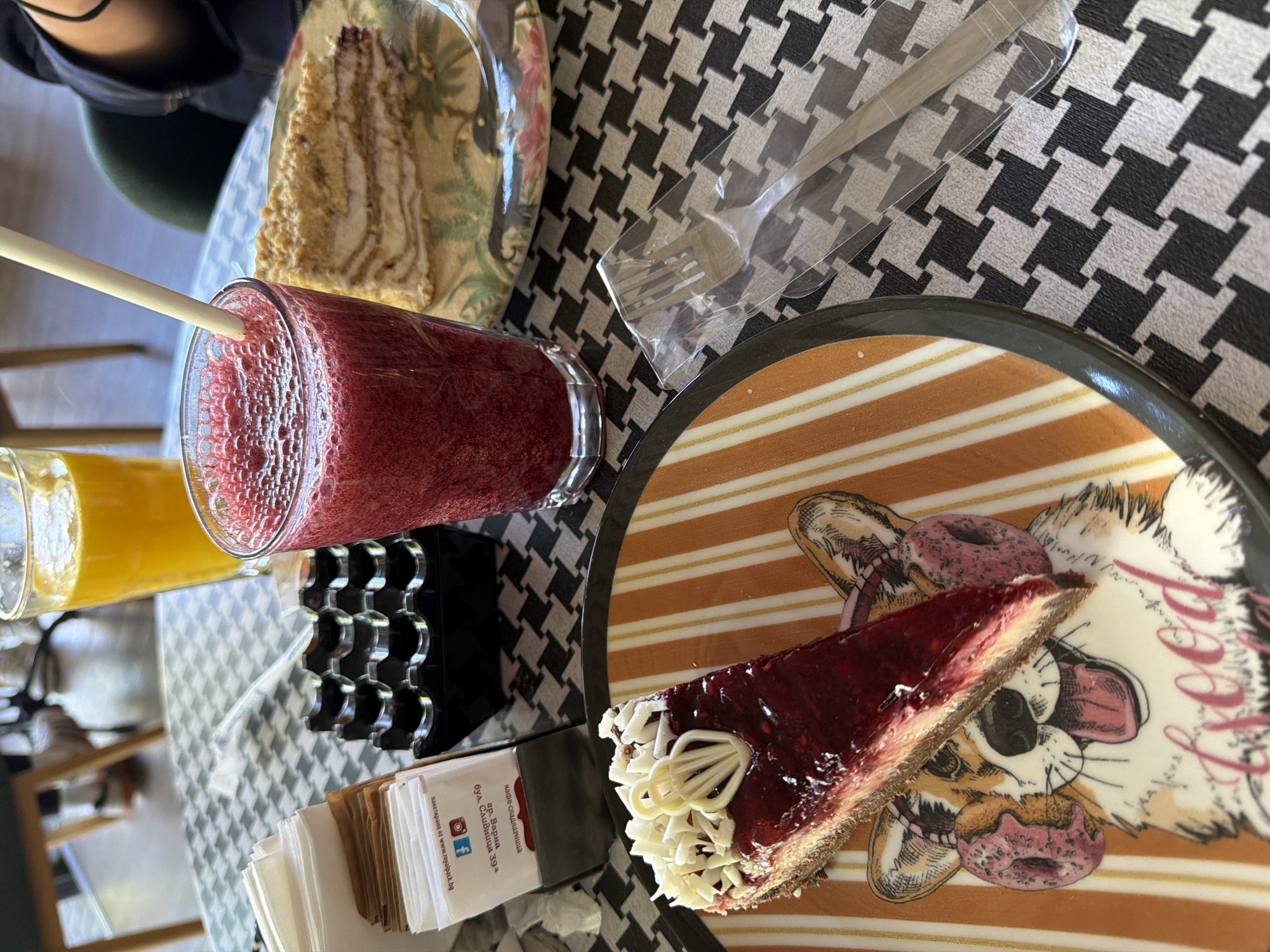 The image shows a table setting with a checkered tablecloth. On the table are a glass of orange juice, a red smoothie with a straw, a slice of cheesecake with red topping and white chocolate swirls on a plate decorated with a cartoon dog and doughnuts. Beside the cheesecake is a stack of promotional cards for a confectionery business at 39 Slivnitsa Boulevard in Varna, Bulgaria. There is also a packeted fork. A piece of cake in a plastic bag sits in the background. A person is partially visible in the upper left.