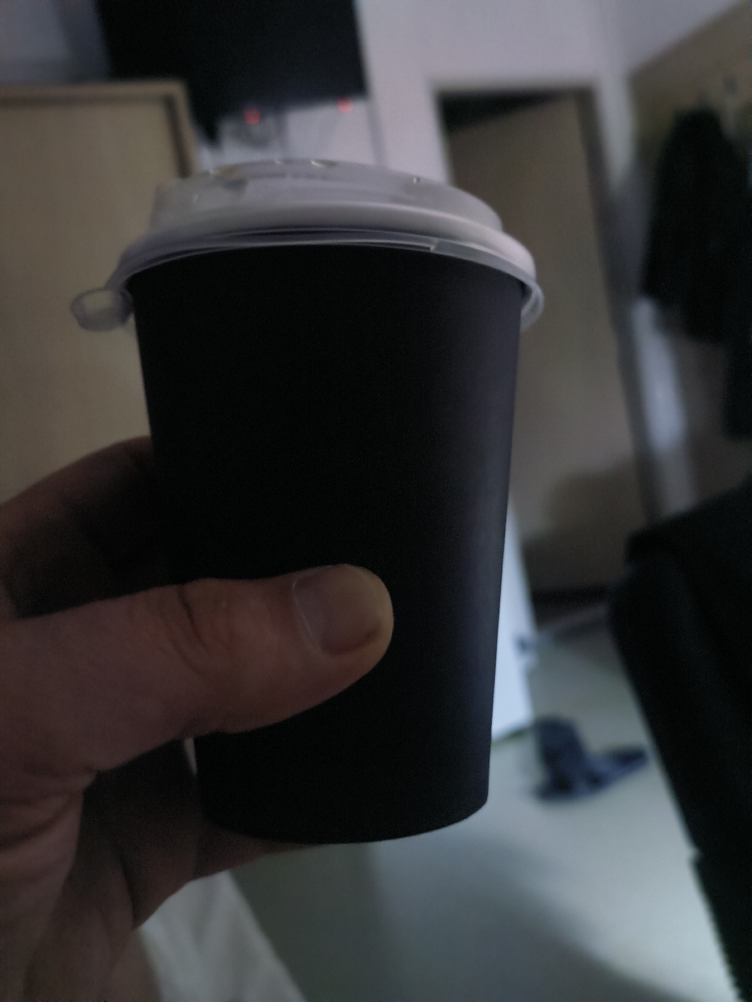 A close-up, blurry, indoor shot shows a hand holding a black disposable coffee cup with a clear plastic lid. The background is out of focus and appears to be a room with a door and a dark object on the floor. The setting is indoors, likely a residential or commercial space, and the image does not provide enough detail to confirm its location in Қосшағыл ауылдық округі, Kazakhstan.