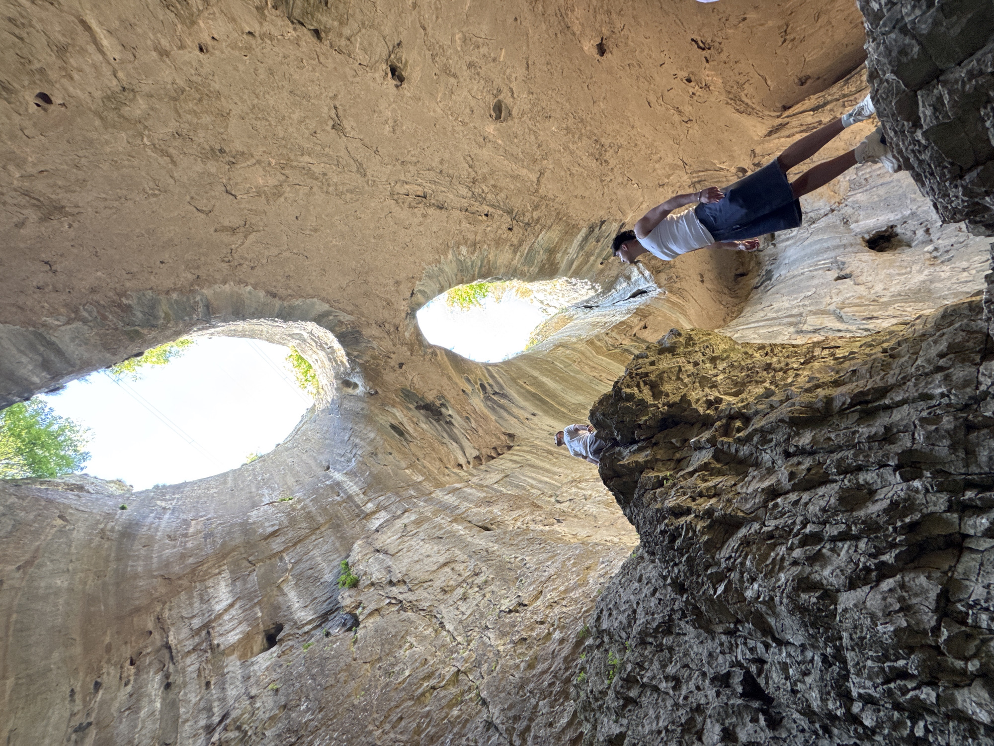 The image shows the Devetashka Cave near Karlukovo, Bulgaria, from an interior, low-angle perspective, looking up. The cave roof has two large openings to the sky, with visible daylight and some tree foliage. A man stands on a rocky ledge at the upper right of the frame. He is wearing a white t-shirt, dark shorts, and white shoes. He appears to be looking down or out from the ledge. The cave walls are made of layered, light-colored rock formations. The foreground rock is darker and more textured.