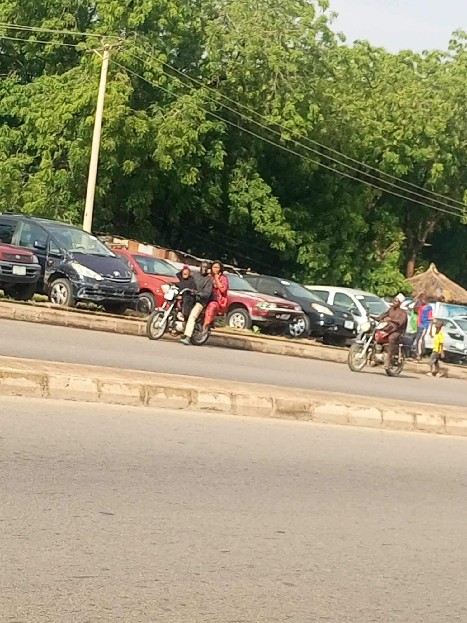 This image captures a street scene in Gombe, Nigeria, with a line of parked vehicles on the left, including a dark blue minivan and red sedans. In the foreground, two motorcycles are visible. The first motorcycle, to the left, is being ridden by two individuals, a person in a dark jacket and possibly black pants, and another person in a pink patterned dress and headscarf. The second motorcycle, further to the right, is ridden by a person in a brown traditional outfit and a white cap. They are observed to be riding in the same direction as the first motorcycle. Behind the parked cars, lush green trees form a dense backdrop, with power lines strung across them and a utility pole on the far left. The ground is paved with asphalt, and a concrete curb runs along the edge of the road. The overall impression is a typical urban street in Gombe, characterized by the presence of vehicles, people on motorcycles, and abundant greenery.