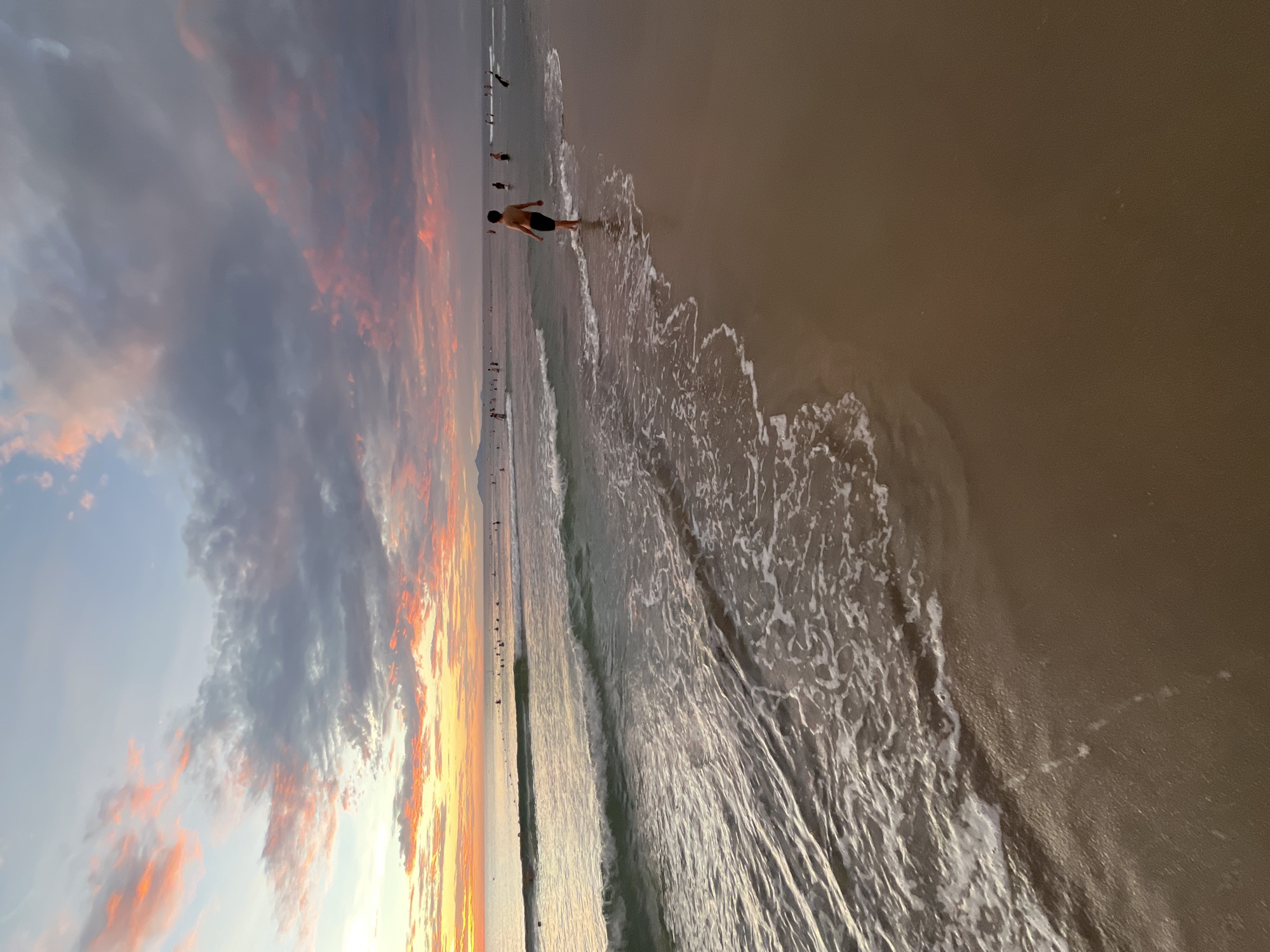 A panoramic landscape photograph captures a serene beach scene at sunset or sunrise in Đà Nẵng, Vietnam. The image, originally rotated, shows a vibrant sky dominating the left side, painted with striking hues of orange, pink, and deep red, blending into lighter blues and grays, interspersed with soft, scattered clouds.
The right side of the frame features the coastline, where gentle waves break onto the sandy shore. White foam from the receding waves creates intricate patterns on the wet sand, which reflects the warm colors of the sky. The sea itself appears calm, stretching into the distance.
Several people are visible along the shoreline. In the midground, a prominent figure, a man in dark swim trunks, stands in the shallow water, facing away from the camera, seemingly observing the horizon. Further down the beach, more smaller figures are scattered, suggesting others are enjoying the evening or morning by the sea.
The overall atmosphere is peaceful and picturesque, highlighting the beauty of the coastal landscape at a golden hour. There is no visible text in the image.