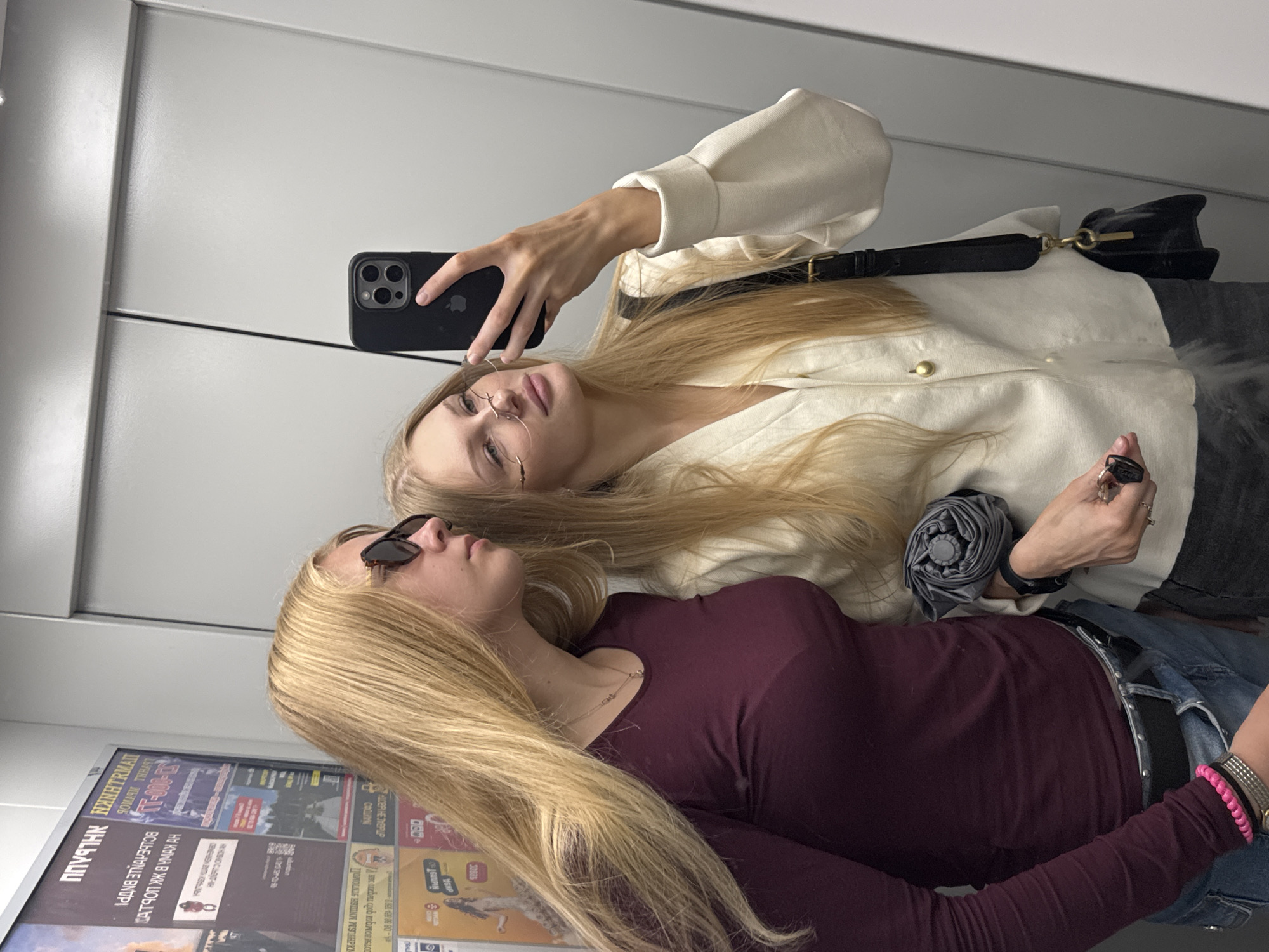 Two young blonde women are taking a selfie in an elevator. The woman on the left is wearing a burgundy long-sleeved shirt and blue jeans, with sunglasses on. The woman on the right is wearing a white jacket and glasses. She is holding a black smartphone and an umbrella. The elevator has grey walls and doors. The background appears to be in the city of Perm, Russia, as indicated by posters on the elevator wall.
