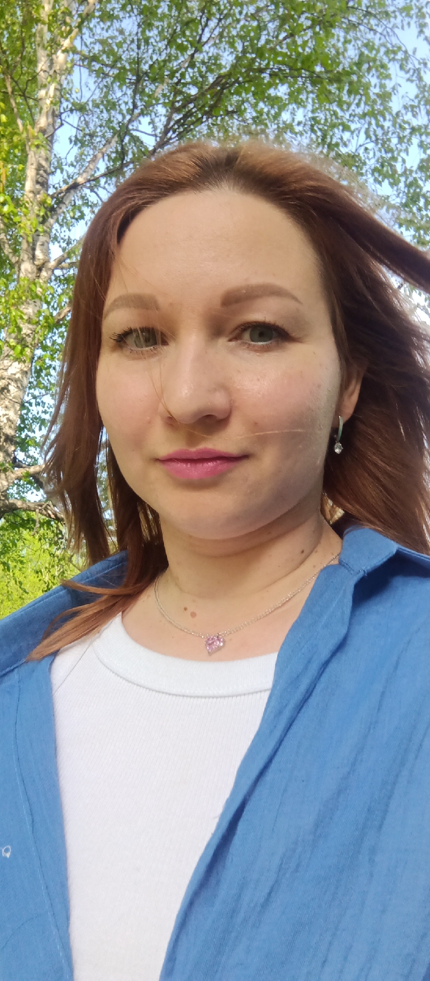 A woman with reddish-brown hair, light skin, and green eyes is seen in a selfie-style close-up outdoors. She is wearing a white t-shirt under a blue shirt. A delicate silver necklace with a pink heart-shaped pendant adorns her neck, and she has small silver earrings. The woman's hair is slightly windswept, with strands across her face. In the background, there is foliage, including birch trees. The location is an outdoor area, likely a park or garden, in the city of Unknown, Unknown.