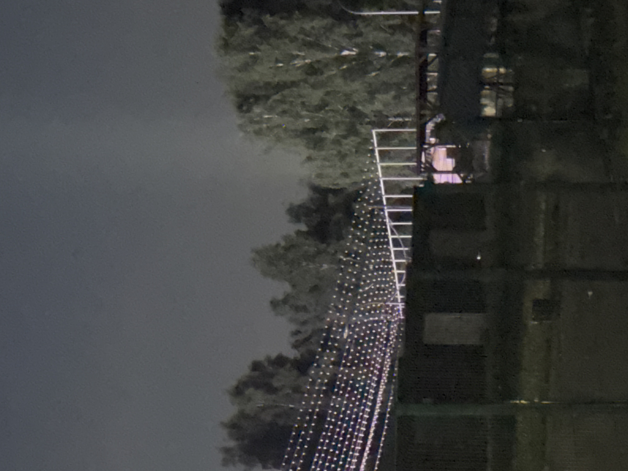 The image shows a nighttime scene in Izhevsk, Russia. A tall structure, possibly a tower, is illuminated with numerous strings of lights, creating a cascading effect. Adjacent to the structure are trees, appearing dark against the night sky. To the right, there's a building or a section of a building, seemingly unlit. The overall environment suggests a public space or park decorated for an event or holiday.