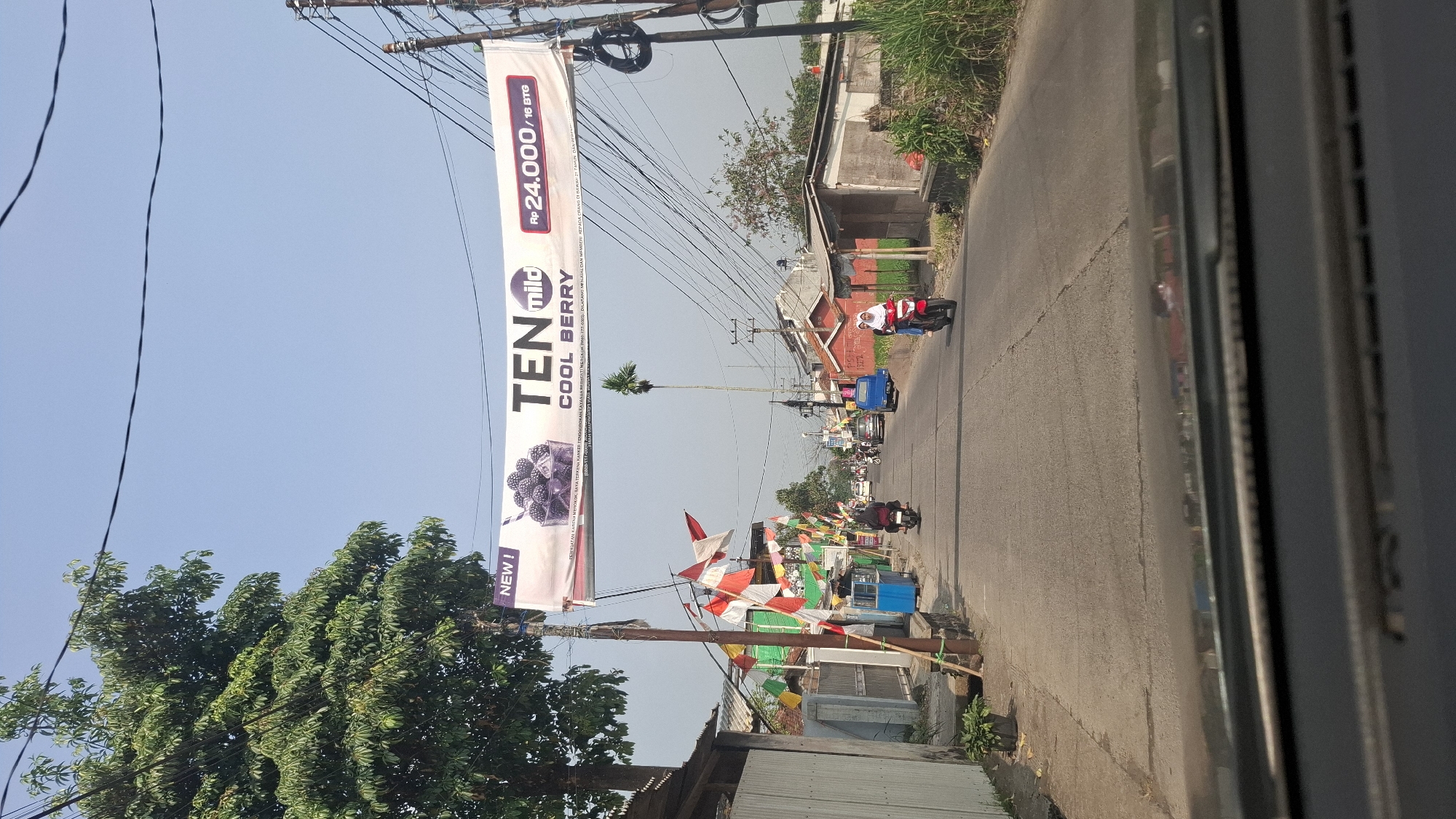 This is a full shot of a street in Cikaroya, Indonesia, captured from the perspective of a vehicle. A banner advertising "TEN mild COOL BERRY" with a price of Rp 24.000/16 BTG is suspended across the road. The banner also contains text in Indonesian, including "NEW!" and a warning about smoking. Several Indonesian flags are displayed on poles along the side of the road. Motorcycles are seen on the road, with riders dressed in casual attire and one rider wearing a hijab. Buildings line both sides of the street, some appearing residential. The sky is clear and blue, and the weather appears to be sunny. The road surface is asphalt and shows some wear. A tall palm tree is visible in the background.