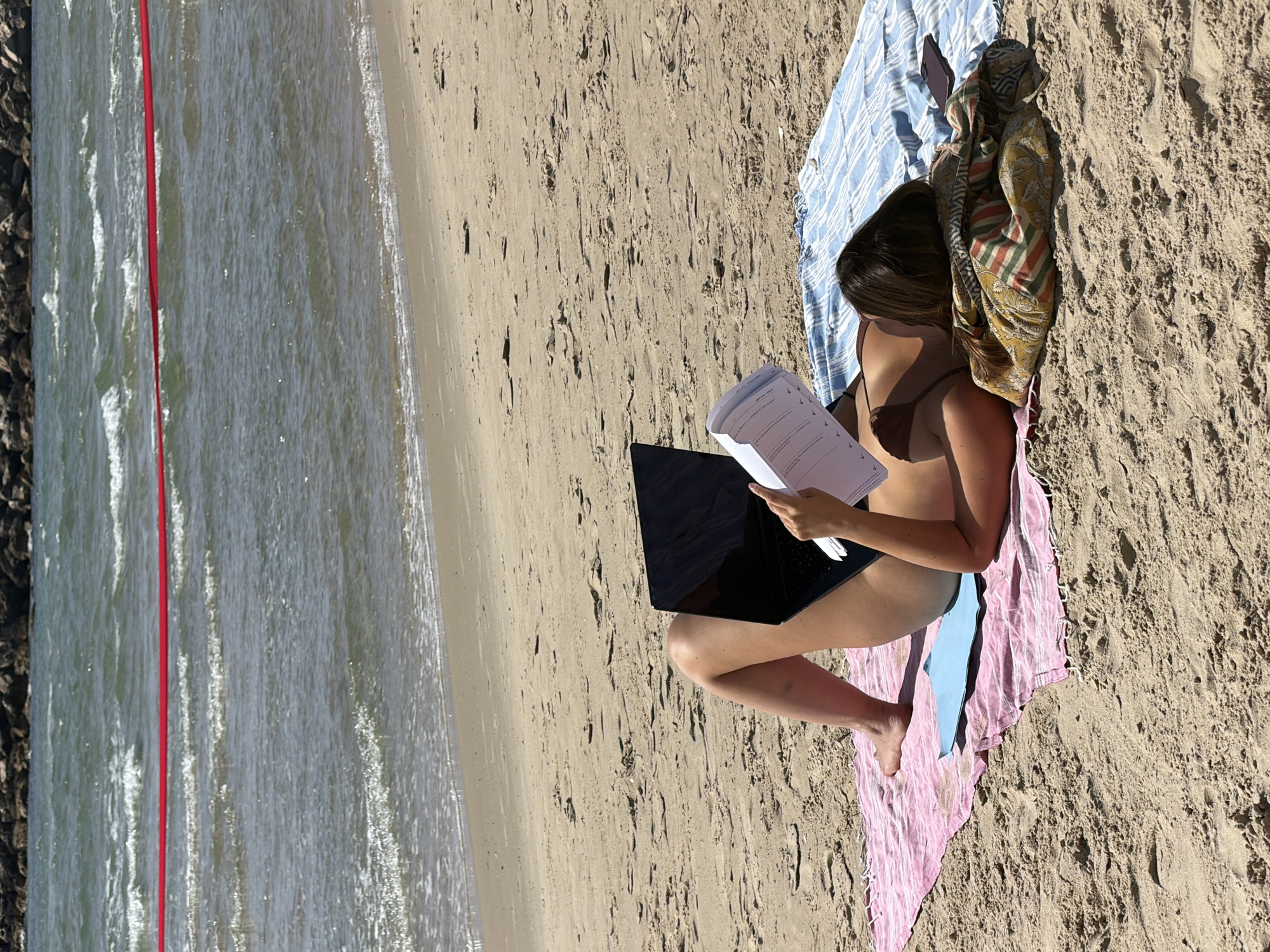 A woman is sitting on a multi-colored towel at a beach, in Tel-Aviv, Israel. She is wearing a brown swimsuit and is using a black laptop while reading a document. The beach is sandy and slopes towards the water, and the weather is sunny.