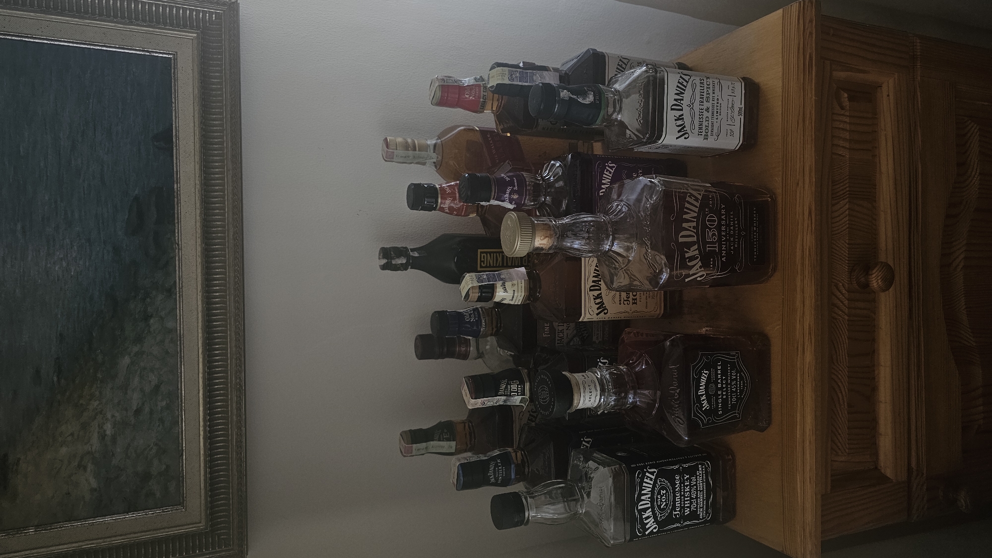 The image depicts a collection of alcoholic beverages, primarily various bottles of Jack Daniel's whiskey, arranged on a wooden surface, likely a piece of furniture. There are also other brands visible, including Johnnie Walker. The bottles are of different sizes and packaging. To the left of the bottles is a framed painting or print. The scene appears to be indoors. There is no information in the image to indicate it is specifically located in Rogachevo, Bulgaria.