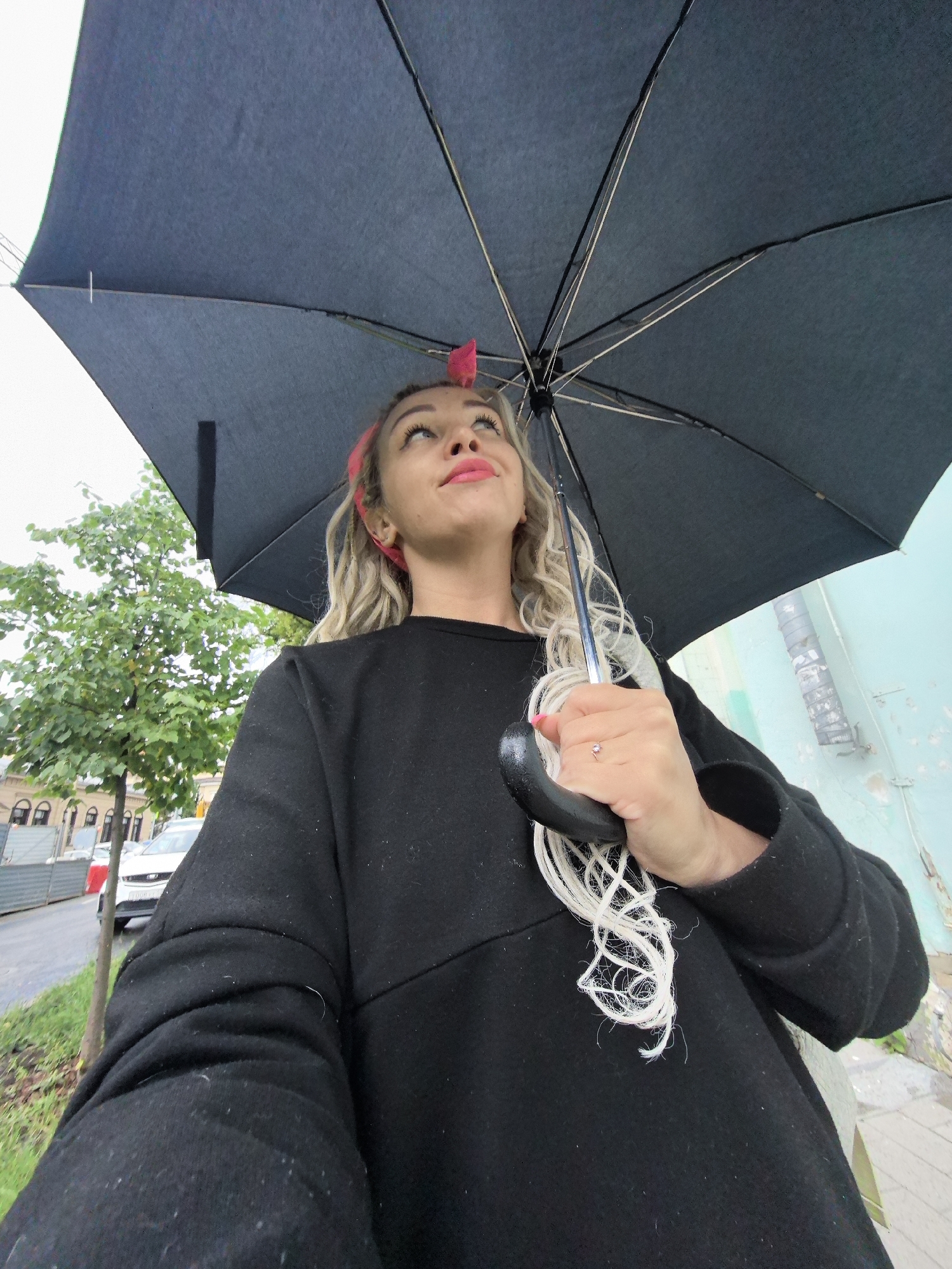A medium shot, low-angle perspective shows a young woman holding a black umbrella overhead. She is looking upwards, with her face framed by blond, curly hair and a bright pink headscarf. She wears a dark, long-sleeved sweater and a small ring with a gemstone on her left hand, which is gripping the umbrella's handle.
The background reveals a city street scene. To the left, a green tree with dense foliage stands beside a sidewalk and a road where a white car is parked. Buildings with arched windows line the street. To the right, a textured, light blue wall with visible pipework and peeling paint suggests an urban setting. The overall lighting indicates an overcast day, possibly with light rain, given the presence of the umbrella. The image is devoid of specific landmarks that would pinpoint it to Nizhny Novgorod, Russia.