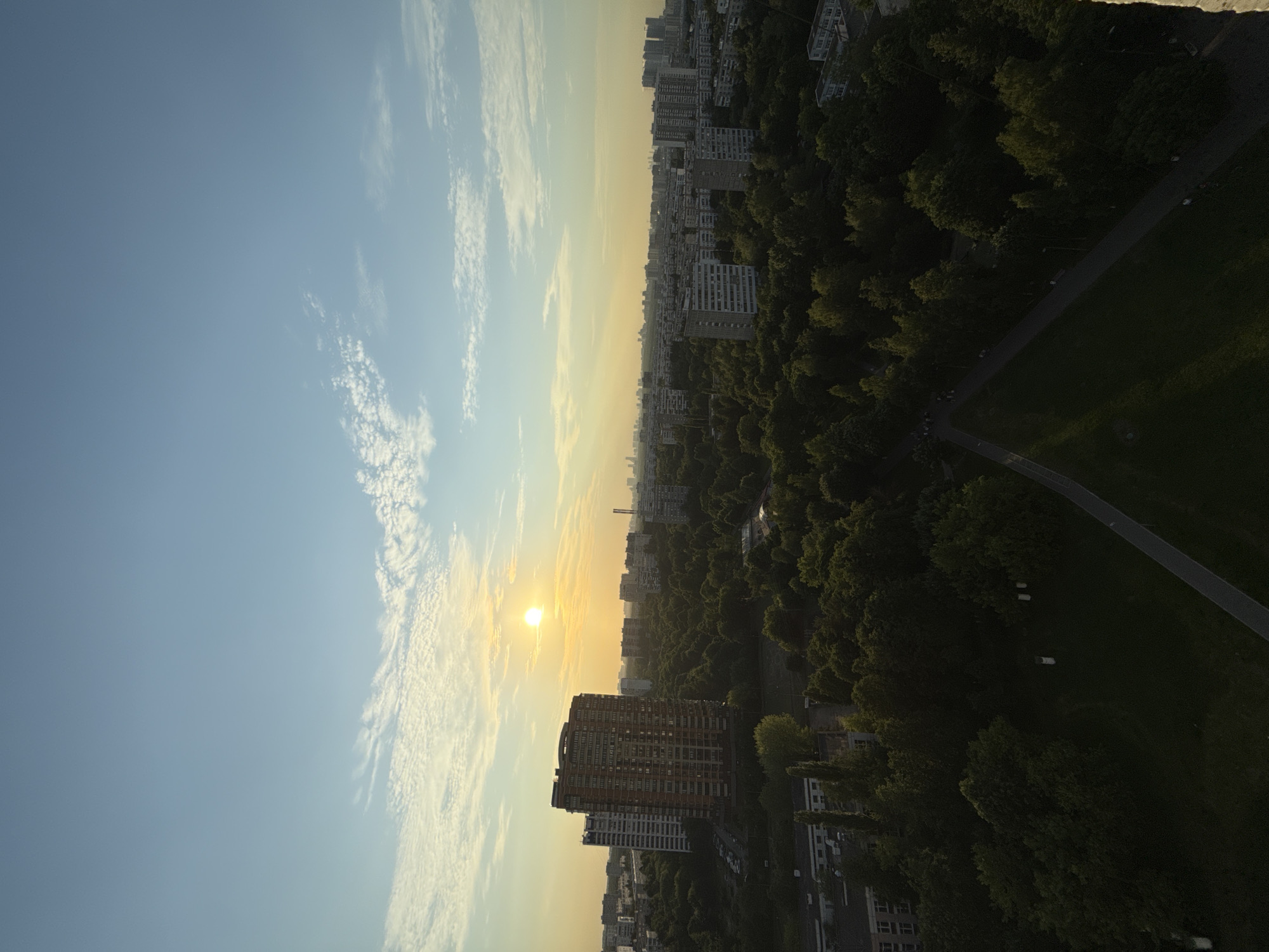 An aerial view captures a city skyline bathed in the warm glow of a setting sun. The sky, a gradient of pale blue and soft yellow, is dotted with wispy clouds. In the foreground, a dense canopy of lush green trees dominates the landscape, interspersed with apartment buildings. A prominent, multi-story brick building stands out, its facade illuminated by the sun. Further in the distance, rows of high-rise buildings stretch across the horizon, silhouetted against the colorful sky. A paved path winds through the park-like area in the foreground. The scene is tranquil, showcasing a harmonious blend of urban development and natural greenery.