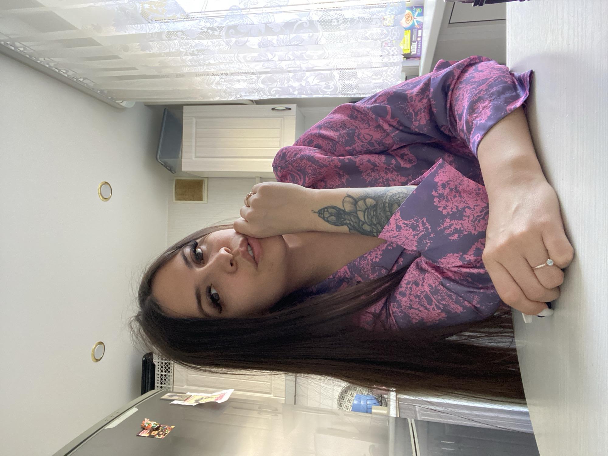 A woman with dark brown hair is seated at a white table in a kitchen. She is wearing a purple and pink patterned robe. Her left hand is at her face and she has rings on both hands. She has a tattoo on her left forearm. The kitchen has white cabinets and a window with sheer curtains. There are magnets on a refrigerator visible in the lower left. The location is likely in Chaikovsky, Russia.