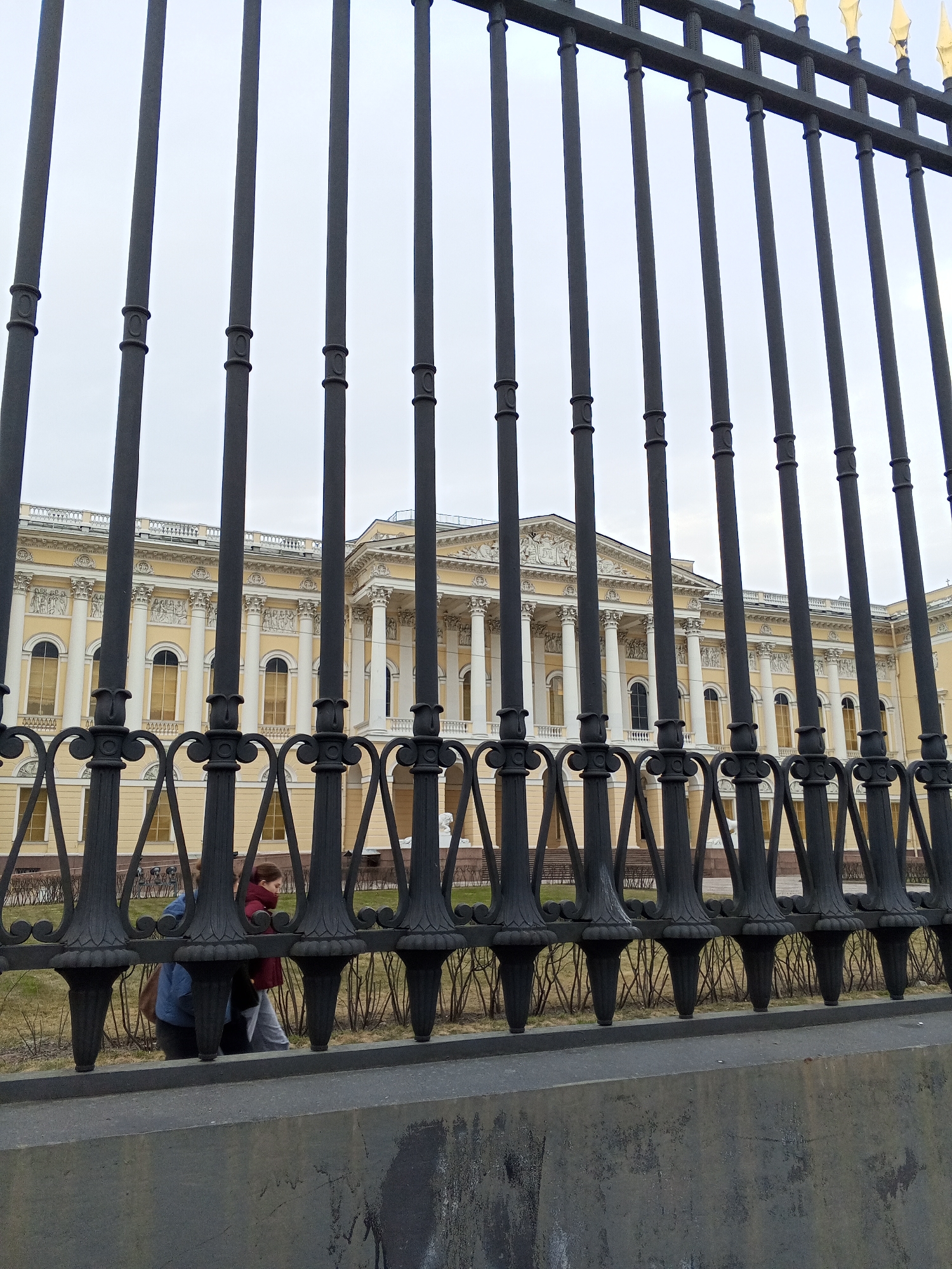 The image shows a view of the Russian Museum in Saint Petersburg, Russia, as seen through a wrought-iron fence. The fence is in the foreground and slightly obscures the view of the museum, which is located in the background. A person is walking behind the fence in the direction from left to right. The sky is overcast.