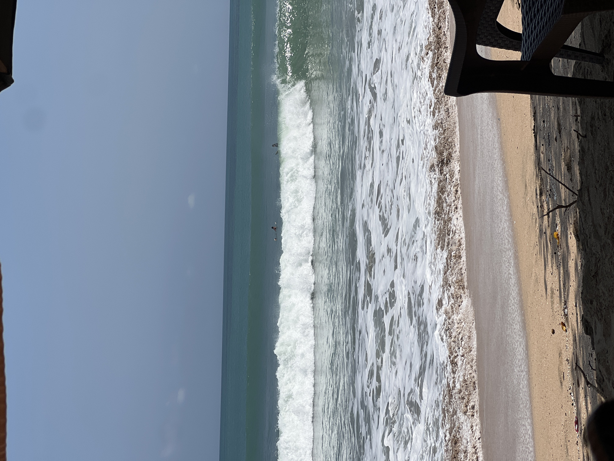 This is an outdoor, eye-level shot of a beach in Kuta, Indonesia, on a clear, bright day. The foreground is dominated by a sandy beach with some debris, including small shells and twigs. A dark brown plastic chair is positioned on the right side of the frame, partially obscuring the view of the sand. The middle ground features the ocean with a large wave breaking. Several surfers are visible in the water, some riding the wave. The background shows the expanse of the ocean and the horizon, with a clear blue sky above. The water is a vibrant turquoise, and the breaking wave is filled with white foam. The overall scene conveys a typical beach day with recreational activities.