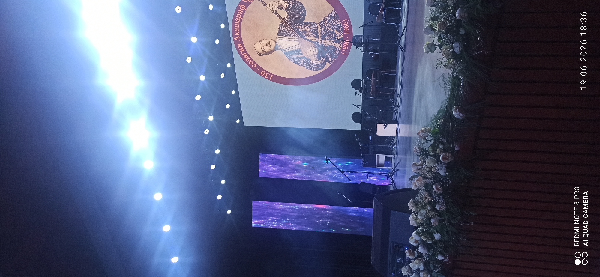 This is a photograph of a stage setup, possibly for a concert or cultural event. The most prominent feature is a large screen displaying a portrait of a man in traditional attire holding a musical instrument, with text surrounding him. The text on the screen reads "130 - солагии Акашариф" and "(1896 - 1966)".
On the stage, there are several microphones set up on stands, suggesting a musical performance. Behind the microphones, there are chairs and what appears to be part of an orchestra. Two large, rectangular screens with a starry, galaxy-like pattern are also visible on the stage, likely serving as backdrops or visual elements.
The scene is illuminated by bright stage lights, creating lens flares and a vibrant, energetic atmosphere. To the right of the stage, there is a floral arrangement, primarily composed of white and cream-colored flowers, adding a touch of elegance to the setting. The background is dark, typical of a theater or performance hall.
In the lower right corner of the image, there is text that reads "19.06.2026 18:36" and "REDMI NOTE 8 PRO AI QUAD CAMERA", indicating the date and time the photo was taken, and the device used. The overall impression is of a formal event being prepared or underway, with a focus on honoring a historical figure.