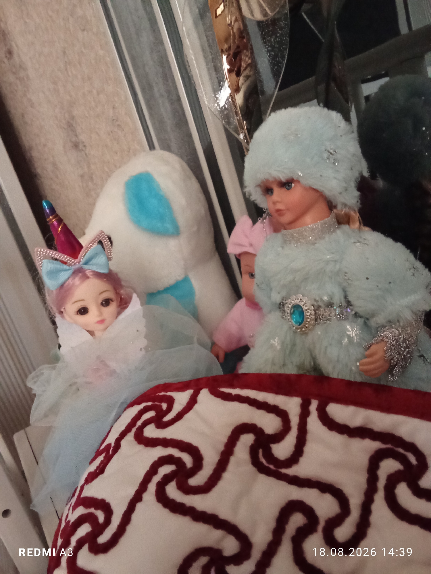 In the city of Khistevarz, Tajikistan, two dolls are prominently displayed in what appears to be a domestic setting. The doll in the foreground on the right is larger and wears a light blue fluffy outfit complete with a matching hat adorned with glitter. It also sports a silver belt with a large blue jewel. To the left of this doll is a smaller doll with pink hair, wearing a white, lacy dress and a unicorn-themed headband with a blue bow. Behind the dolls, a white plush unicorn toy with blue accents is visible, leaning against a striped background that suggests wallpaper or a textile. The scene is framed by a red and white patterned textile in the foreground, possibly a cushion or blanket. The lighting is soft and diffused.