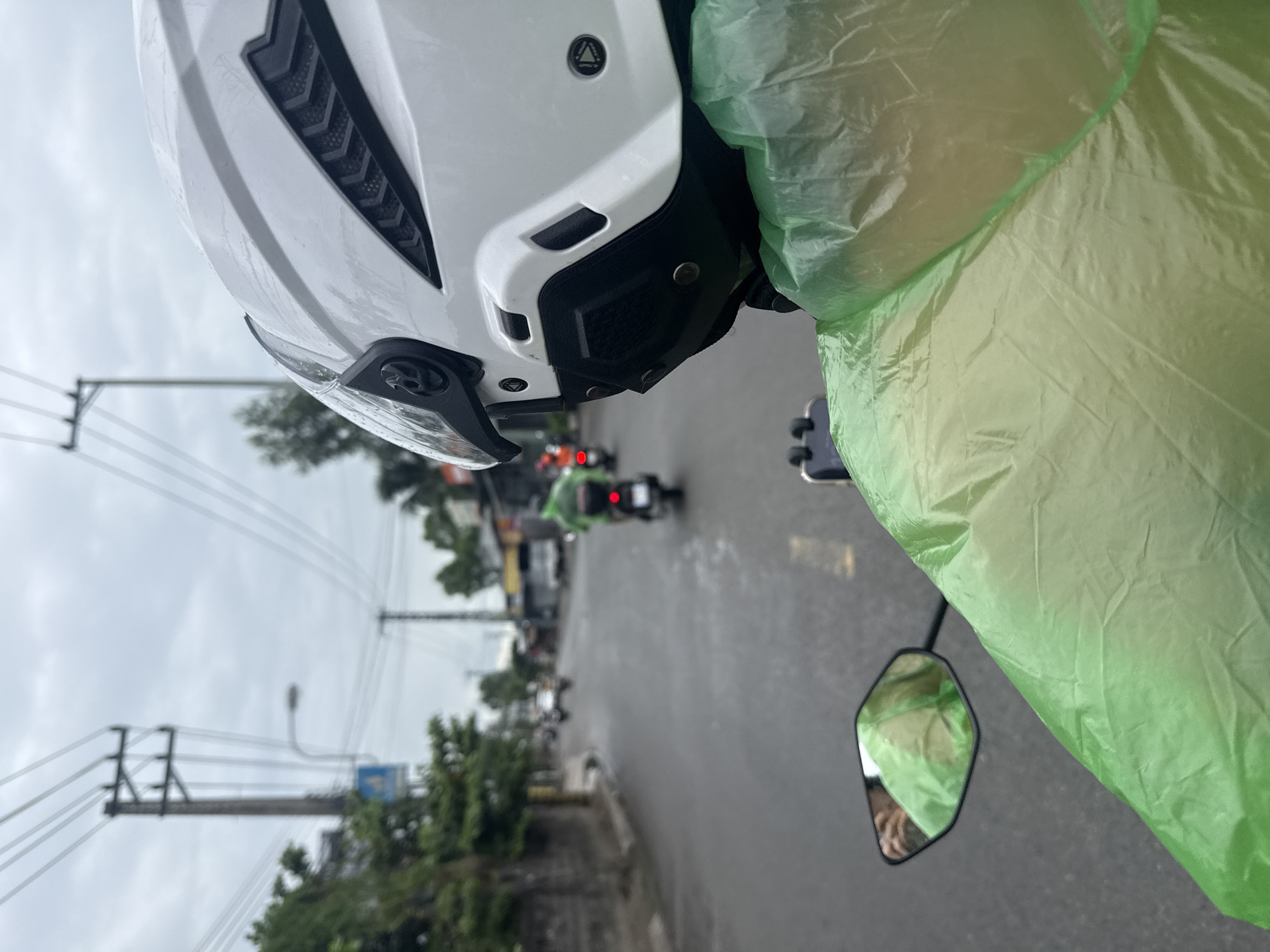 The image depicts a first-person perspective from a motorcycle in Nha Trang, Vietnam. The rider, wearing a white helmet and a green translucent raincoat, is looking ahead at a wet, paved road. Several other motorcycles are visible ahead, some with riders wearing similar green raincoats. Trees and buildings line the sides of the road. Power lines and utility poles stretch across the overcast sky. A mirror on the left side of the motorcycle shows a reflection of the rider, also wearing the green raincoat. The scene suggests travel during or after rain.