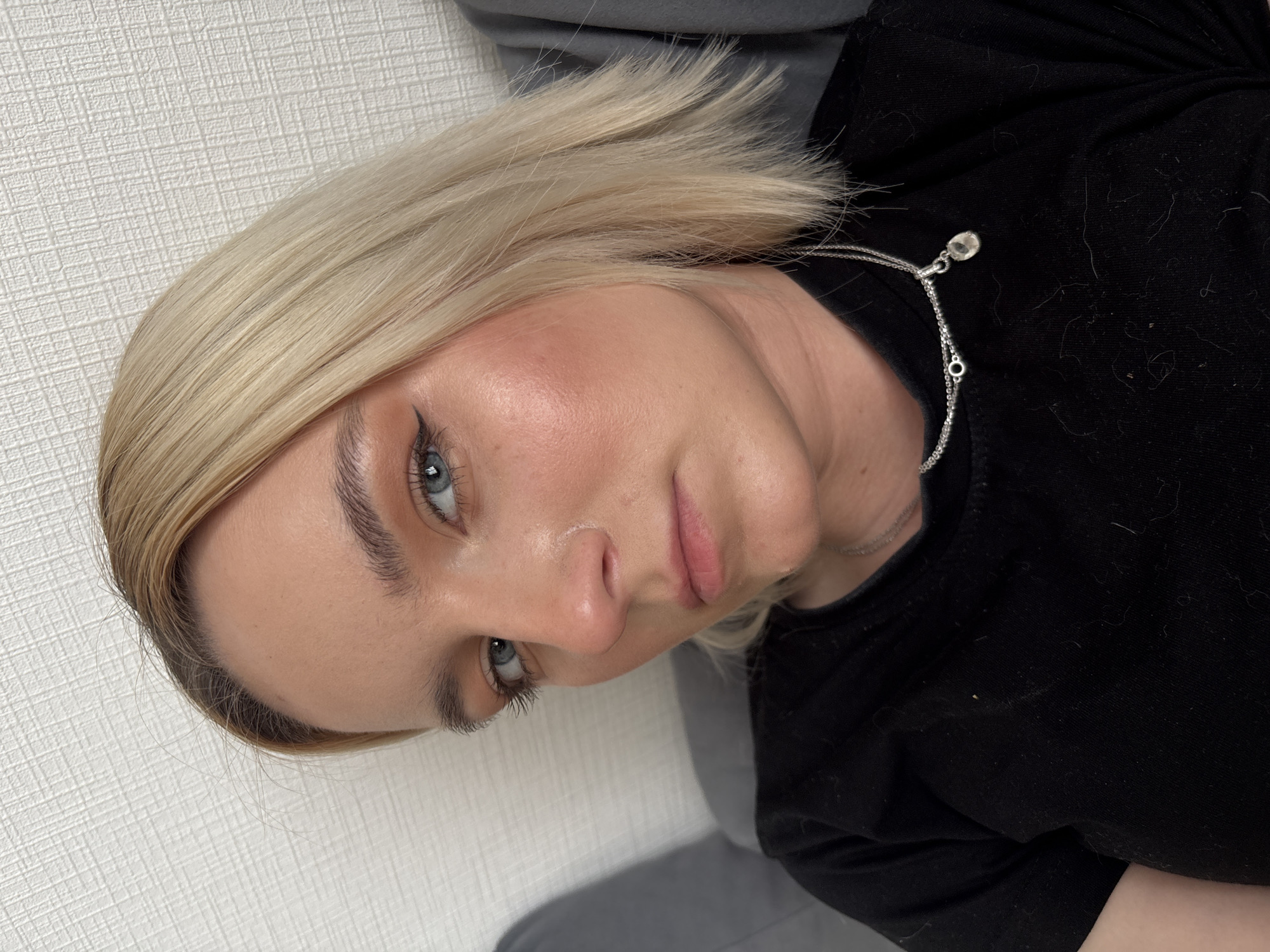 A close-up portrait photograph depicts a fair-skinned woman with blond hair and blue eyes. She is wearing a black shirt and a silver necklace. The background consists of a white wall on the left and a gray surface at the bottom. The photo does not provide information about the location in Vsevolozhsk, Russia, or any specific actions or interactions. The photograph is taken at an angle, with the woman's face slightly tilted.