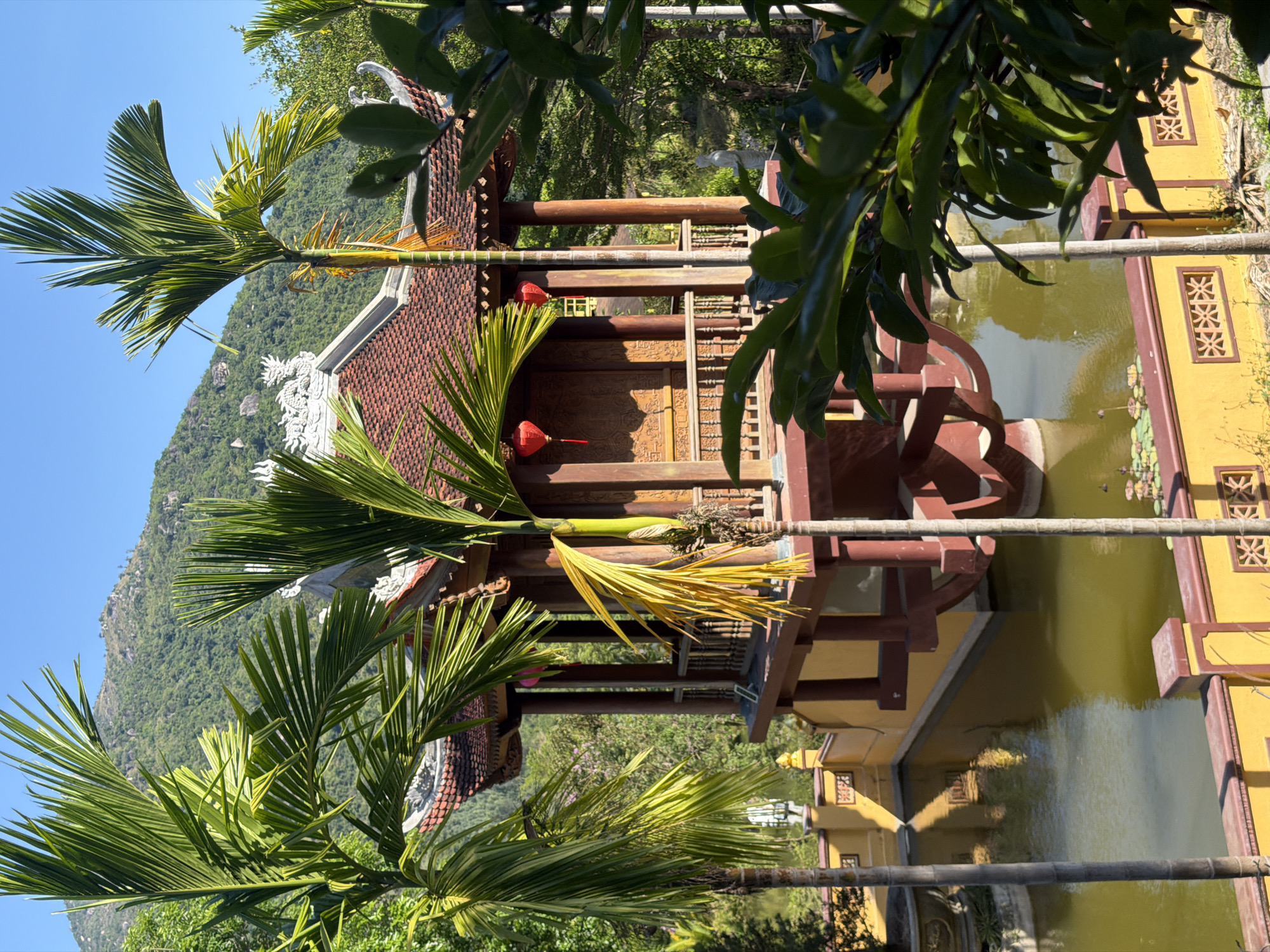 The image depicts a structure built over water, likely a pond or lake, with a traditional Vietnamese architectural style, including a tiled roof and wooden supports. Red lanterns hang from the structure. The building is painted in a combination of brown and yellow hues. In the background, a green mountainous terrain under a clear blue sky is visible. Palm trees are prominent in the foreground, adding a tropical element to the scene. The image suggests a location in Nam Nha Trang Ward, Vietnam.