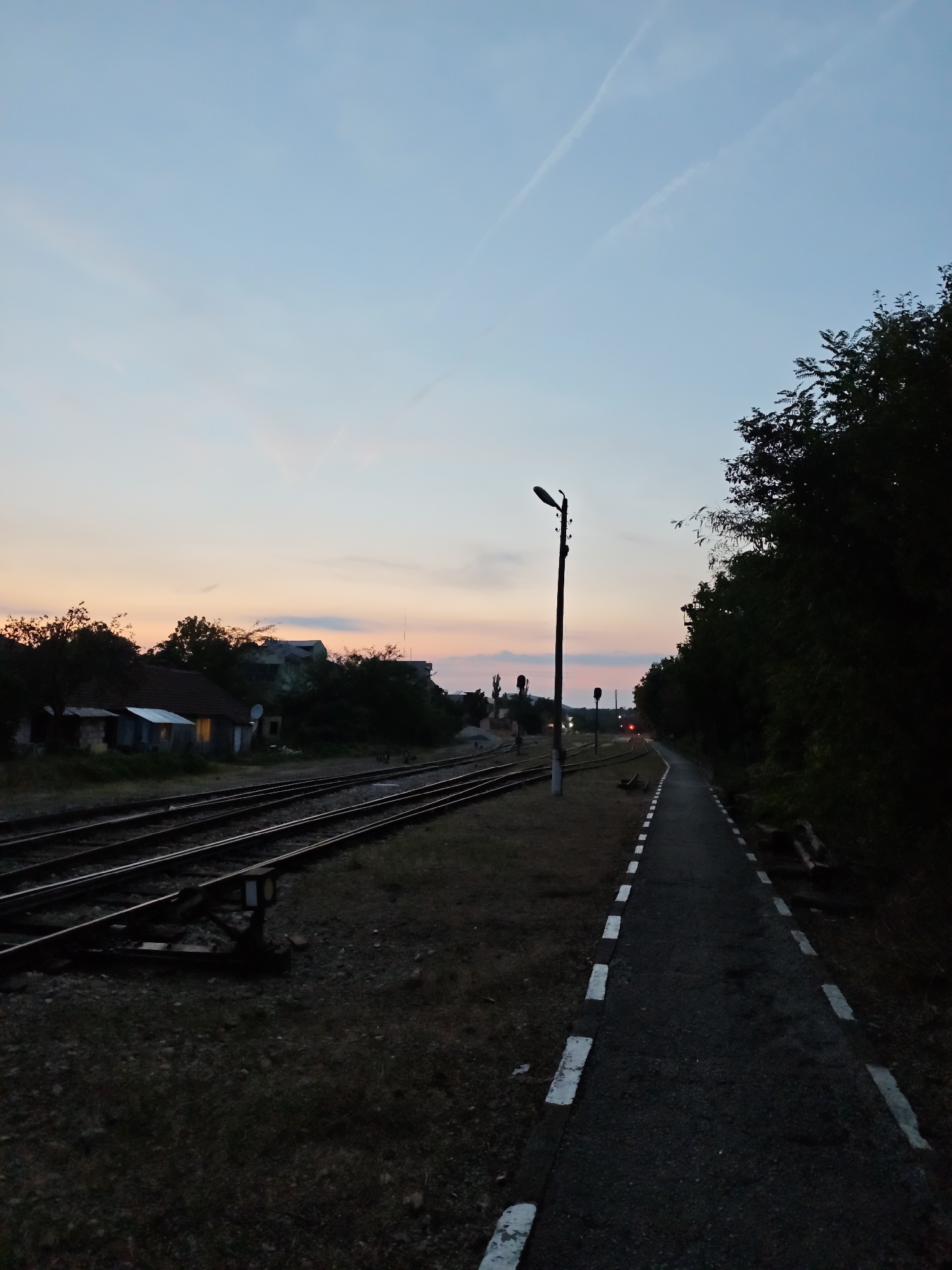 The image depicts a railway track and platform at dusk. The sky is a soft gradient of blue and pink, with faint contrails visible. On the left, multiple railway tracks run parallel, with a small, rustic building visible in the background. A single lamppost stands on the platform to the right, casting a faint light. The platform itself is paved with asphalt and bordered by white stripes. Dense trees line the right side of the platform, creating a dark, shaded area. The scene appears to be a quiet, possibly rural, railway station. Without further information or context, it is not possible to definitively place this scene in Lovech, Bulgaria.
