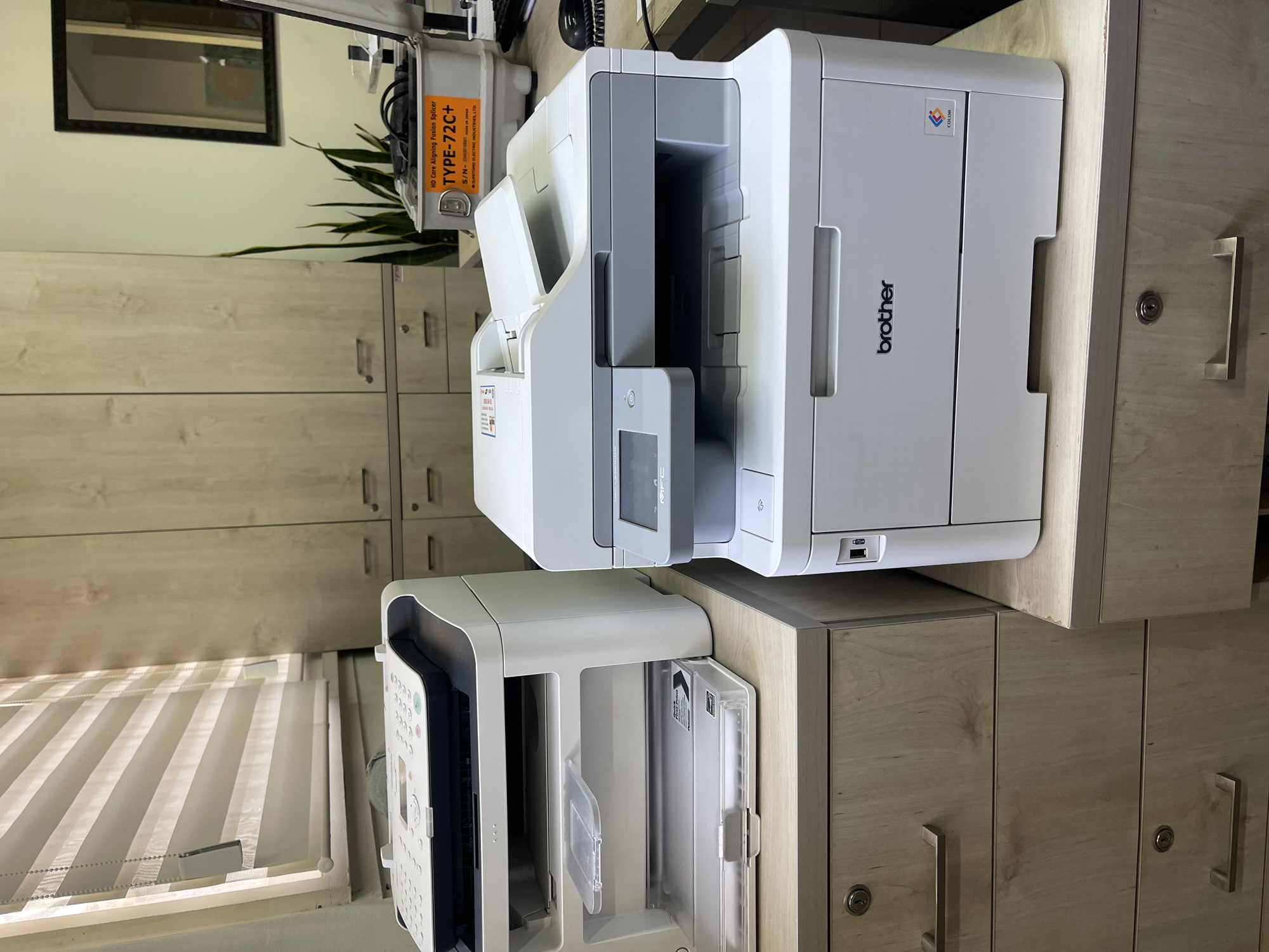 The image shows two printers on a wooden desk in an office. The printer on the left appears to be a fax machine and printer combo, white with a black top. The printer on the right is a white Brother printer, with a control panel and a paper output tray. Behind the printers are light-colored wooden cabinets. A window with blinds is visible on the left, and a plant is seen behind the cabinets. A piece of equipment labeled "TYPE-72C+" is also visible on the desk behind the printers. The scene depicts an office environment with office equipment and furniture. The image does not provide any specific visual information to indicate its location in Tel-Aviv, Israel.