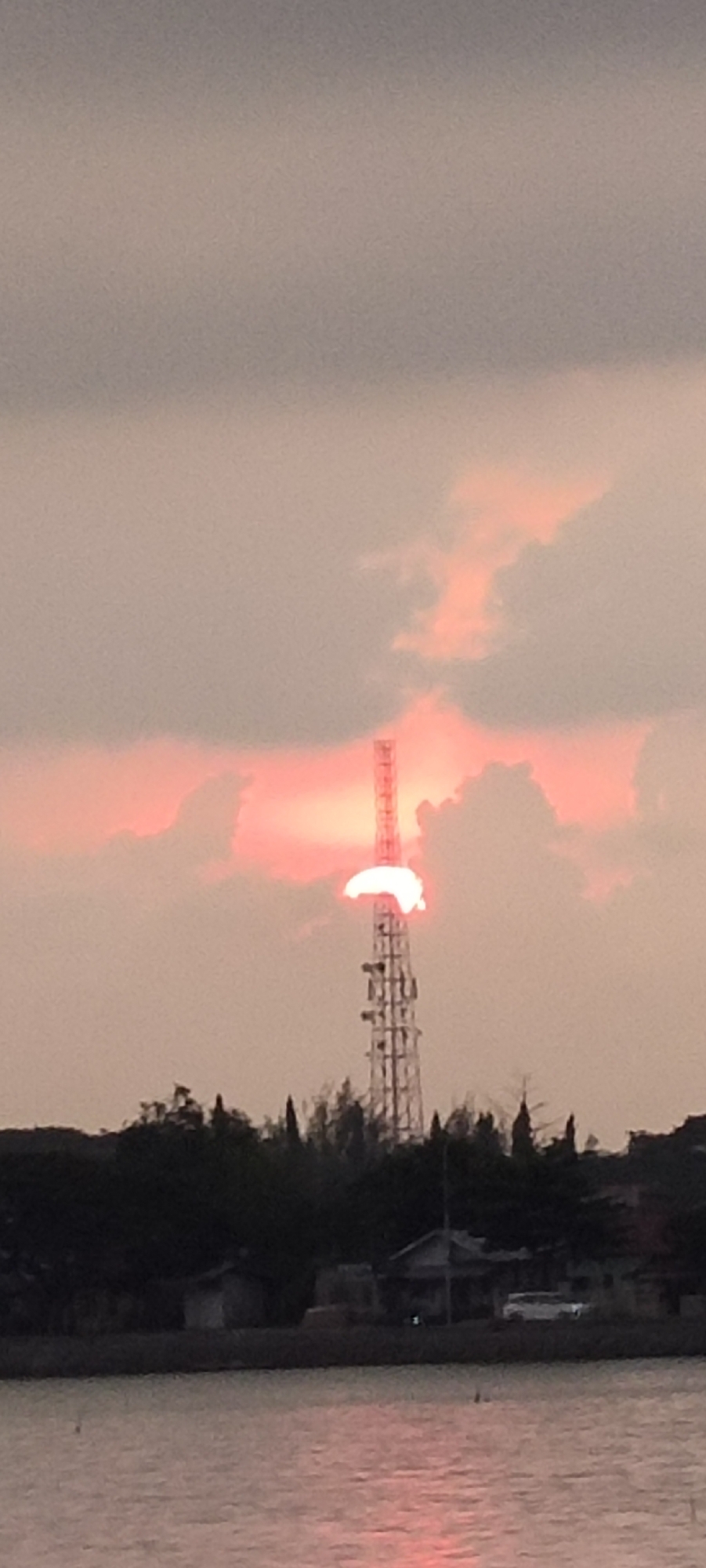The image captures a sunset over a body of water, likely the sea or a large river, in Lhokseumawe, Indonesia. A tall telecommunications tower stands prominently in the middle ground, with the setting sun partially obscured behind it, casting a warm, reddish glow through the clouds. The sky is overcast with soft pink and grey clouds. In the foreground, the water reflects the colors of the sunset. Behind the tower, a line of dark trees and vegetation is visible, with some low-lying buildings and a white vehicle partially illuminated. The overall scene depicts a serene, twilight atmosphere in a coastal or riverside urban setting.