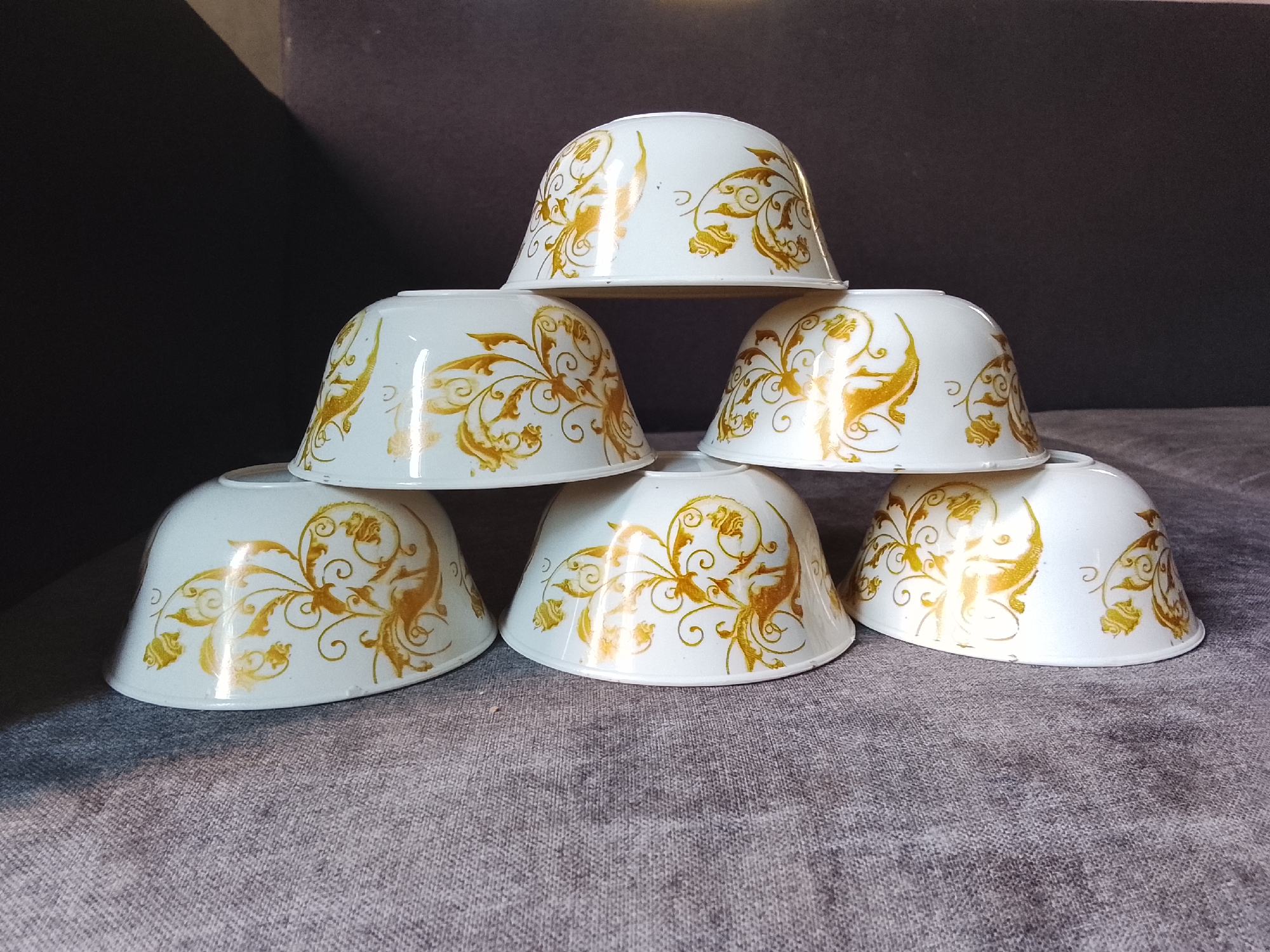 Six white ceramic bowls with gold ornate patterns are stacked in a pyramid formation on a textured grey surface. The bowls are arranged with three in the bottom layer, two in the middle layer, and one at the top. The background is a dark, out-of-focus surface, possibly a couch or wall. The image is well-lit, highlighting the glossy finish of the bowls. The scene depicts a still life arrangement of tableware. There are no people, animals, or any indications of the city of Lubuk Puding, Indonesia.