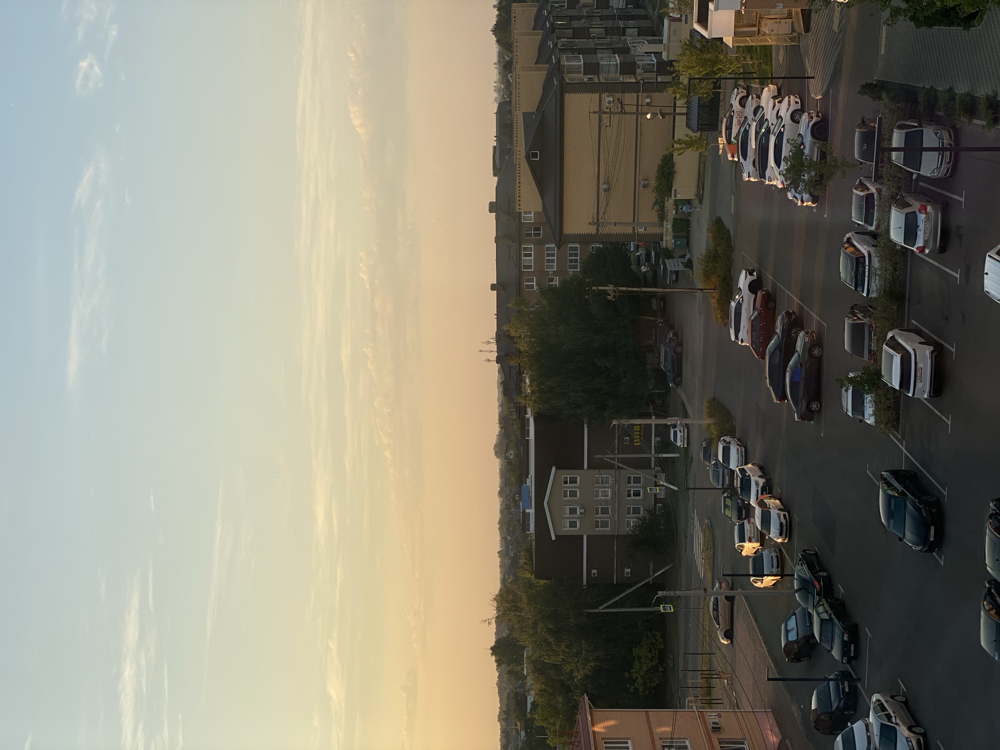 The image depicts a street scene in Prigorodnyi, Russia, during what appears to be sunset. The sky is a gradient of pale blue and warm orange hues, with thin, wispy clouds. Below the sky, a cluster of multi-story residential buildings and a lower-rise commercial building are visible. The commercial building has a sign with Cyrillic lettering.
A paved parking lot occupies the foreground and mid-ground, filled with numerous parked cars of various makes and models, predominantly in darker colors like black, gray, and white. Some cars are in designated parking spots, while others are parked along the roadside. Streetlights are present in the parking lot.
Trees and other greenery are interspersed between the buildings and along the edges of the scene, suggesting a suburban or semi-urban environment. Several utility poles and wires are visible, typical of Russian infrastructure. One white car with a license plate featuring Cyrillic characters is prominently featured in the lower right of the image. The lighting suggests late afternoon or early evening, casting long shadows across the parking lot. No people are clearly identifiable in the scene.