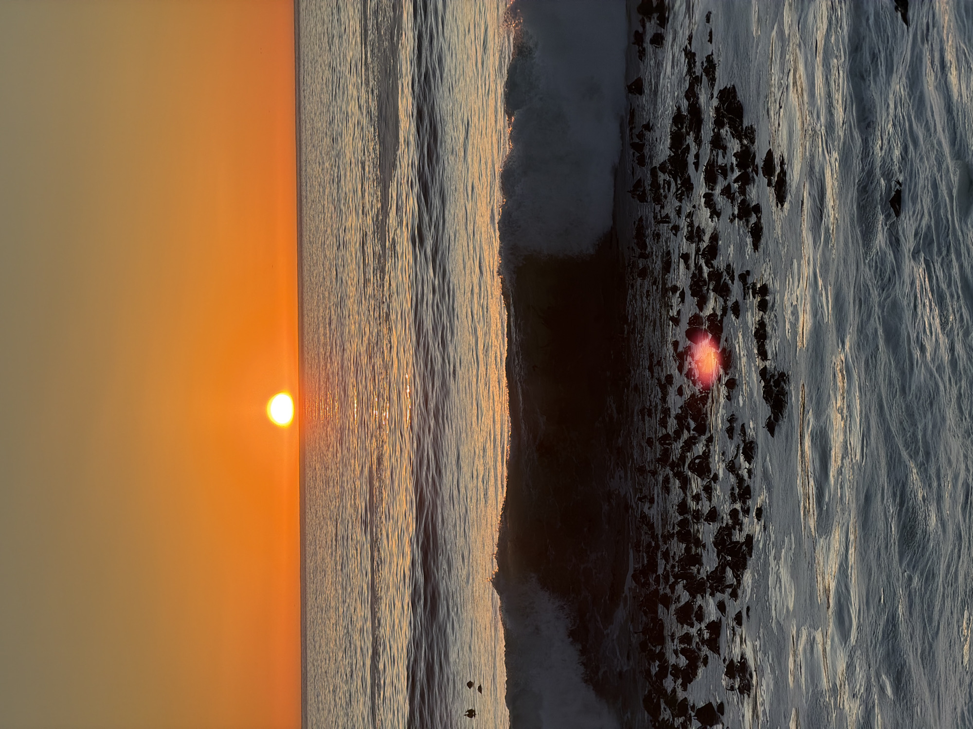 This image captures a sunset over the ocean at a rocky coastline, likely in the Camps Bay or Clifton area of Cape Town, South Africa, given the typical rocky formations and the clear view of the sun setting over the Atlantic. The sun, a bright orange orb, is low on the horizon, casting a warm glow across the sky and reflecting on the water. The ocean is calm with gentle waves lapping against the shore. In the foreground, dark, seaweed-covered rocks emerge from the water, creating a textured and rugged shoreline. Small waves are breaking over these rocks, with white foam contrasting against the dark, wet surfaces. A sun flare is visible as a bright red-orange spot on the water, to the left of the center of the frame, in the foreground. In the mid-ground, a larger wave is cresting, its white foam catching the light. A few dark, bird-like figures are visible in the distance, standing on rocks or swimming in the water. The sky transitions from a vibrant orange near the horizon to a paler yellow and then a light brown towards the top of the frame.