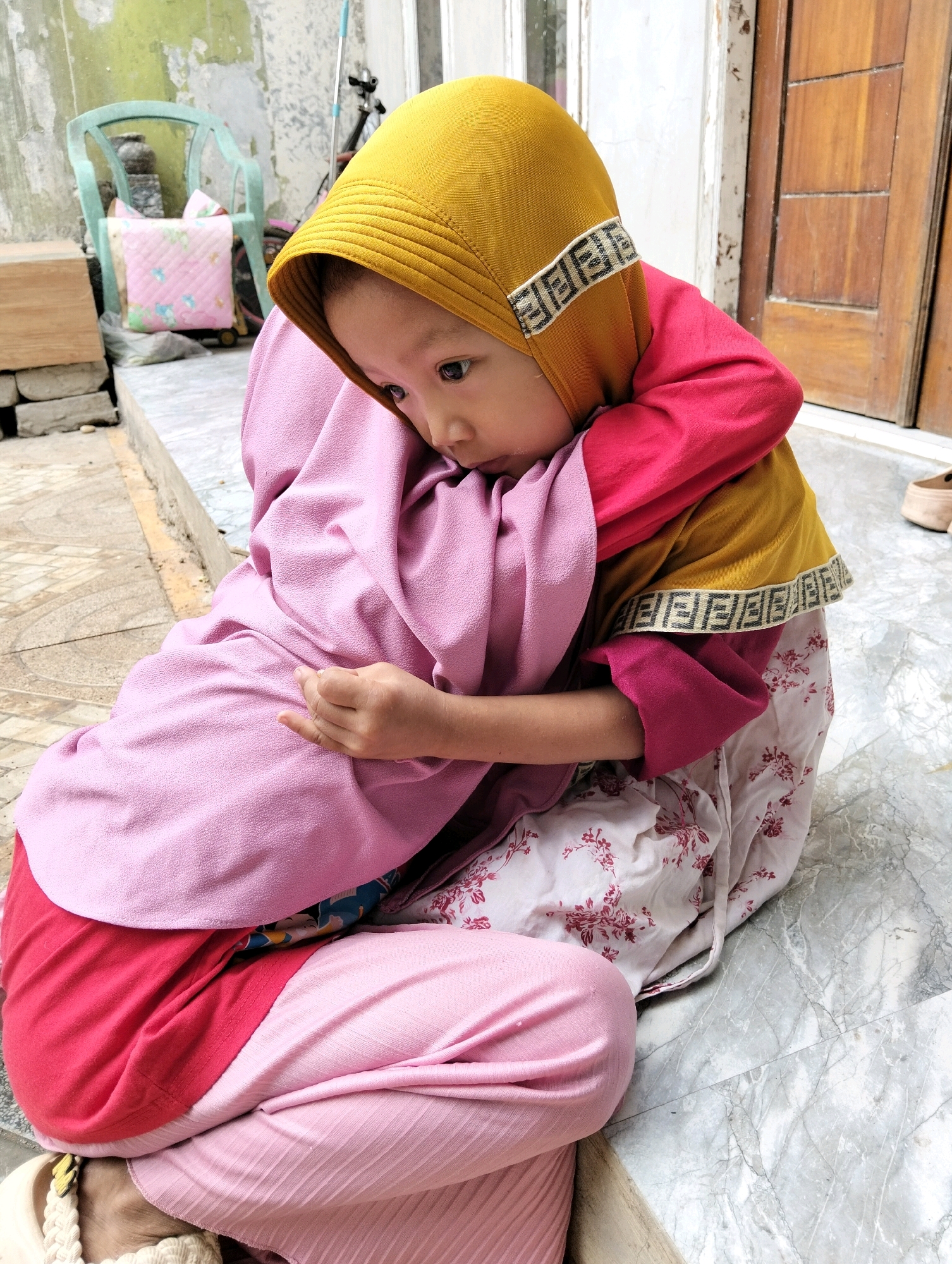 In Cileungsi, Indonesia, two children are embracing outdoors. The foreground features a young child wearing a yellow hijab with a decorative band and a pink shirt, being hugged by another child partially visible and draped in a pink garment. The children are seated on a tiled floor, with a pink and white floral skirt visible on the child in the foreground. In the background, a weathered wall, a green chair with a pink cushion, and wooden doors are present. The environment appears to be an outdoor residential area.
