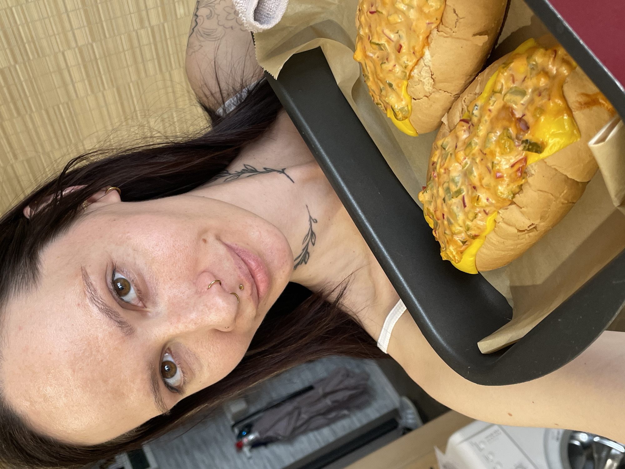 A woman with light skin, brown hair, and multiple piercings in her nose is holding a baking tray containing two bread rolls topped with cheese and a relish-like spread. The woman has tattoos on her neck and arm. The tray is lined with baking paper. The background shows a kitchen counter and appliances. The location is unknown, and there is no information indicating Izhevsk, Russia.