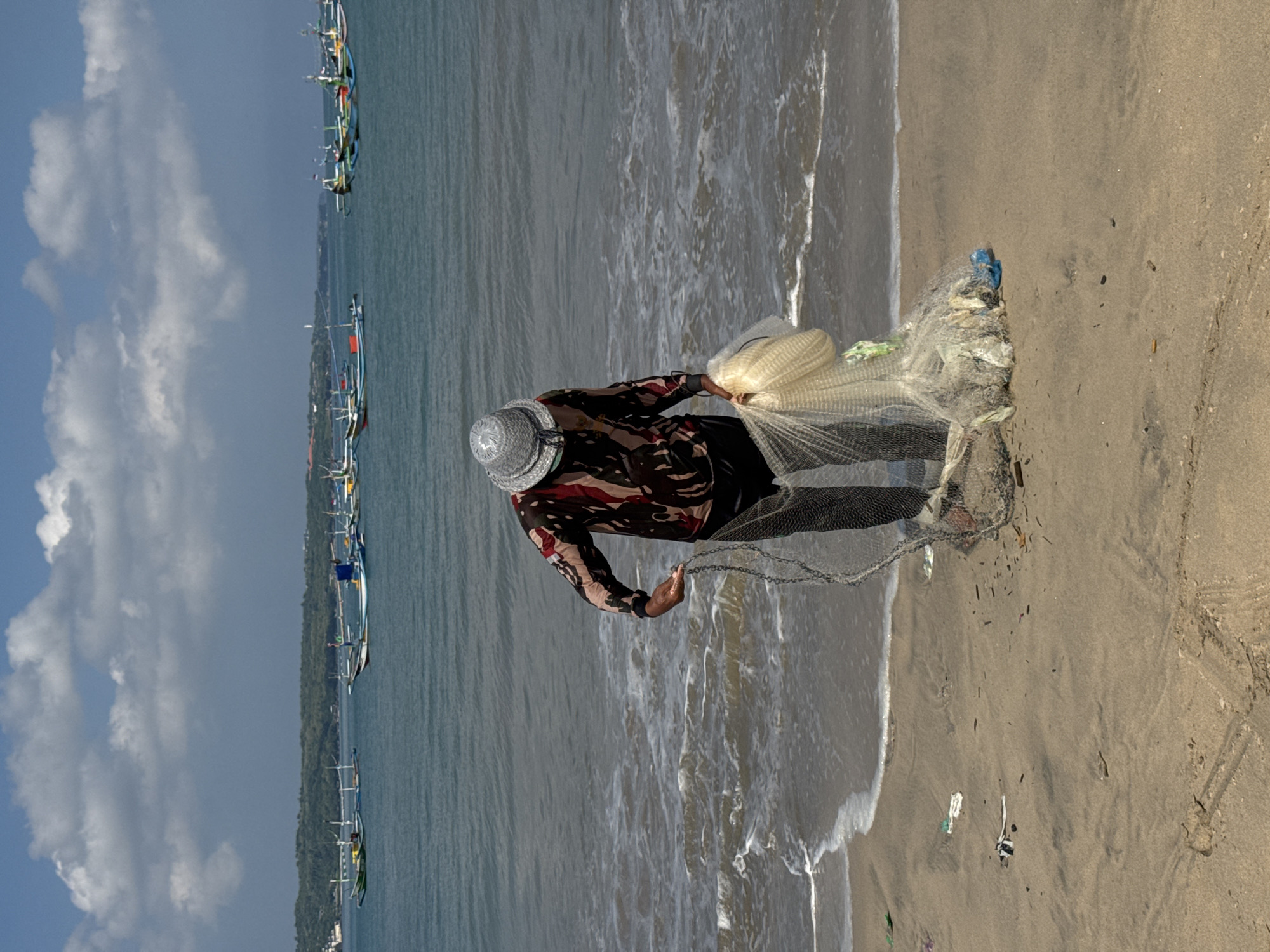 The image, when correctly oriented, depicts a fisherman on a sandy beach at Kuta, Indonesia, preparing a cast net. The person is seen from an elevated perspective, standing at the water's edge, wearing a patterned long-sleeved shirt and a light-colored, possibly woven, hat. They are holding a large, fine-mesh white cast net, part of which lies on the wet sand.
In the background, the sea stretches out under a partly cloudy sky with large white clouds. Numerous traditional Balinese fishing boats, known as jukungs, with their distinctive outriggers, are anchored or floating in the water, extending along the coastline into the distance. A green, tree-covered landmass or island is visible on the distant horizon. The sand closer to the water is wet from the gentle waves, while the higher sand is drier. Small pieces of debris, including plastic and other refuse, are visible scattered on the sand near the water line, particularly around the fishing net. The scene captures a daytime activity, likely showing the preparation for or execution of fishing with a cast net. No visible text is present in the image.