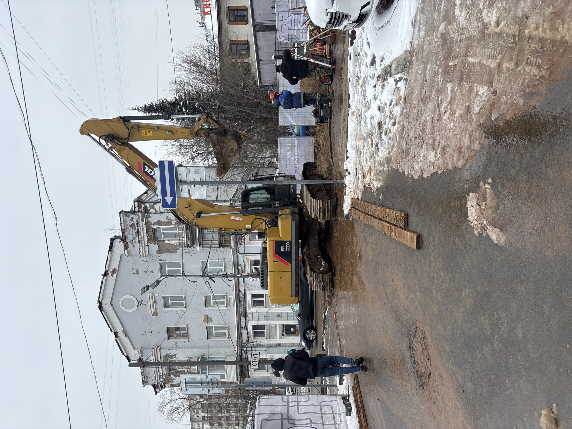 The image depicts a construction site in Perm, Russia, identifiable by Cyrillic signage in the background and the style of the architecture. A yellow excavator is the primary subject, positioned on the street. The excavator is actively digging a trench. There are at least three workers present at the site. Two workers are seen near the trench itself. A pedestrian wearing a dark jacket and jeans is walking on the side. Adjacent to the construction area is a building with peeling paint and multiple windows. Power lines are strung overhead, adding to the urban environment. Patches of dirty snow and slush are present along the side of the road. Two planks are positioned on the road.