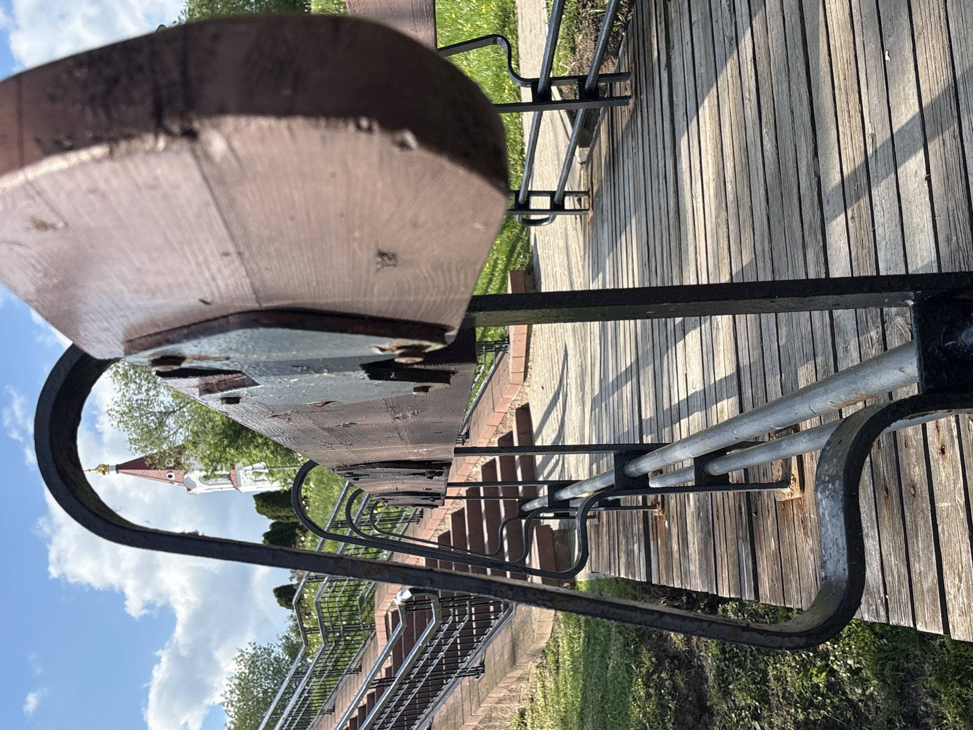 The image is taken from a low perspective, possibly at bench-level, in Minsk, Belarus. The foreground features a worn wooden bench with a metal frame. In the background, there is a partially obscured view of a building with a distinctive red roof and gold dome, suggesting a church or cathedral. The scene also encompasses a wooden walkway, grassy areas, and metal railings leading to steps. The sky is visible, with white clouds suggesting a bright day.