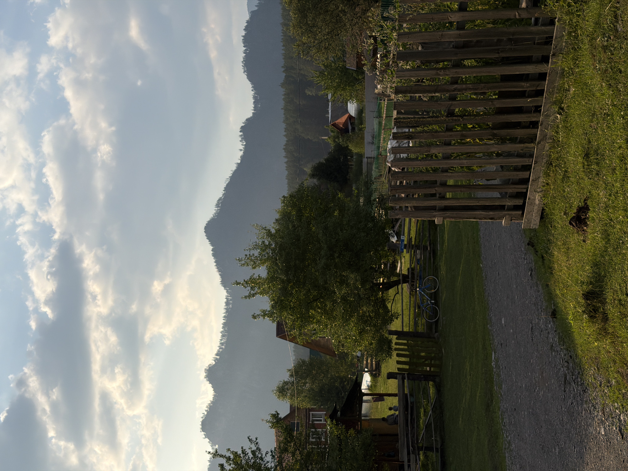 A photograph captures a rural scene in the village of Askat, Russia, depicting a small settlement nestled against a backdrop of forested mountains. The sky above is filled with scattered white clouds, partially obscuring the sun, which casts a warm, soft light on the landscape.
In the foreground, a gravel path leads into a grassy yard. To the right, a rustic wooden fence, constructed from vertical planks, borders a garden area. Beyond this fence, a lush green lawn stretches out.
The middle ground reveals several wooden houses with red and brown roofs, suggesting traditional Russian architecture. A large, leafy green tree stands prominently in the center, its branches reaching towards the sky. To the left of the tree, a blue bicycle is propped up against a wooden fence. Two figures, appearing to be adults, are seated beneath a covered wooden structure attached to one of the houses. A smaller figure, possibly a child, is standing nearby. Another fence, also made of wood, encloses a portion of the yard.
In the background, dense green trees form a dark line across the landscape, separating the village from the imposing, hazy mountains that dominate the horizon. The mountains are covered in a light mist, softening their contours and giving the scene a serene, almost ethereal quality. The overall impression is one of tranquility and a simpler way of life, characteristic of a remote village setting.