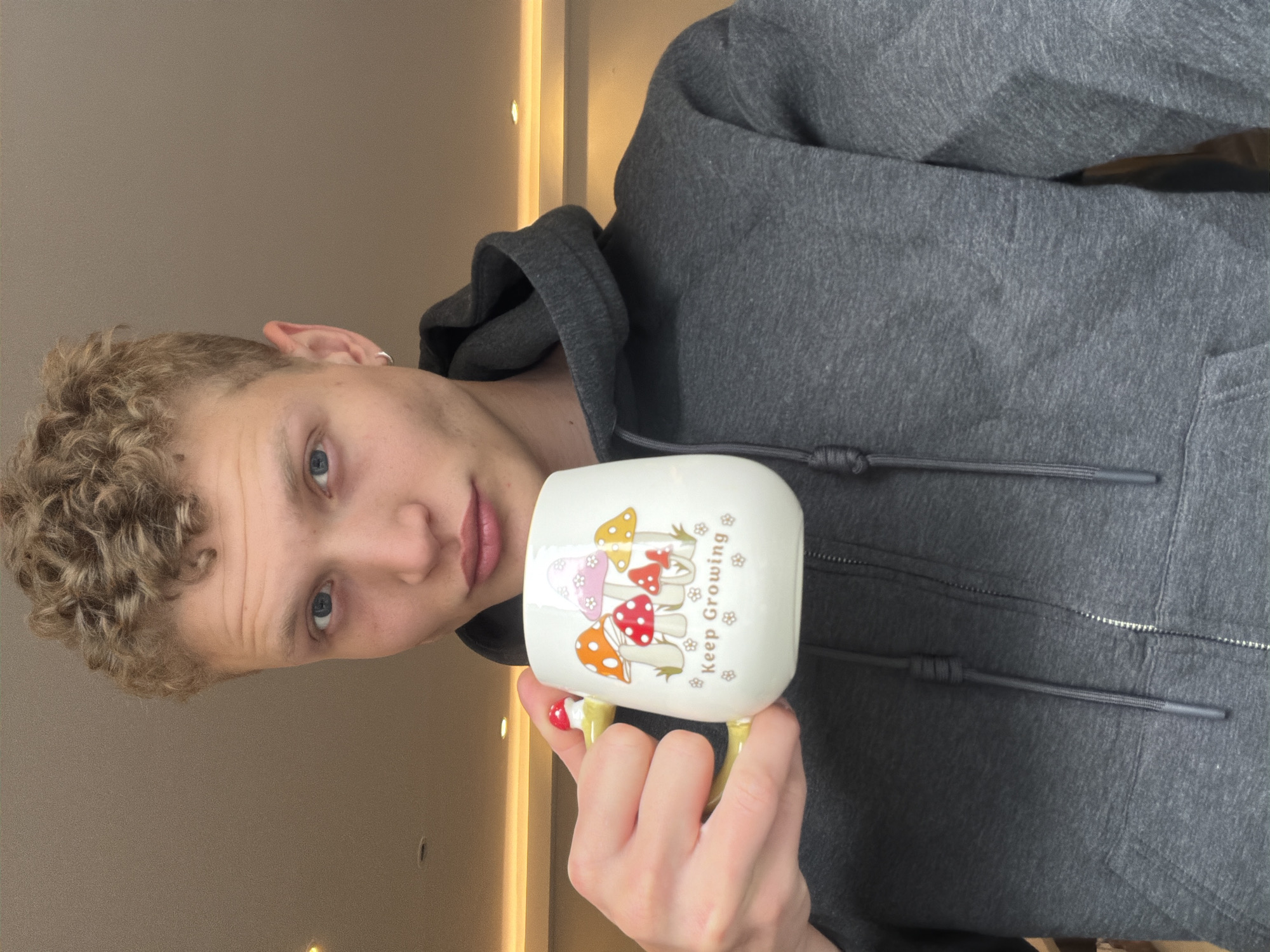 In this medium shot, a fair-skinned, blonde-haired individual with blue eyes is holding a white mug with a mushroom design and the words "Keep Growing" printed on it. The individual is wearing a dark grey hooded sweatshirt. The background consists of a neutral-colored wall and ceiling, with some lighting fixtures visible. No specific location in London, United Kingdom is depicted.