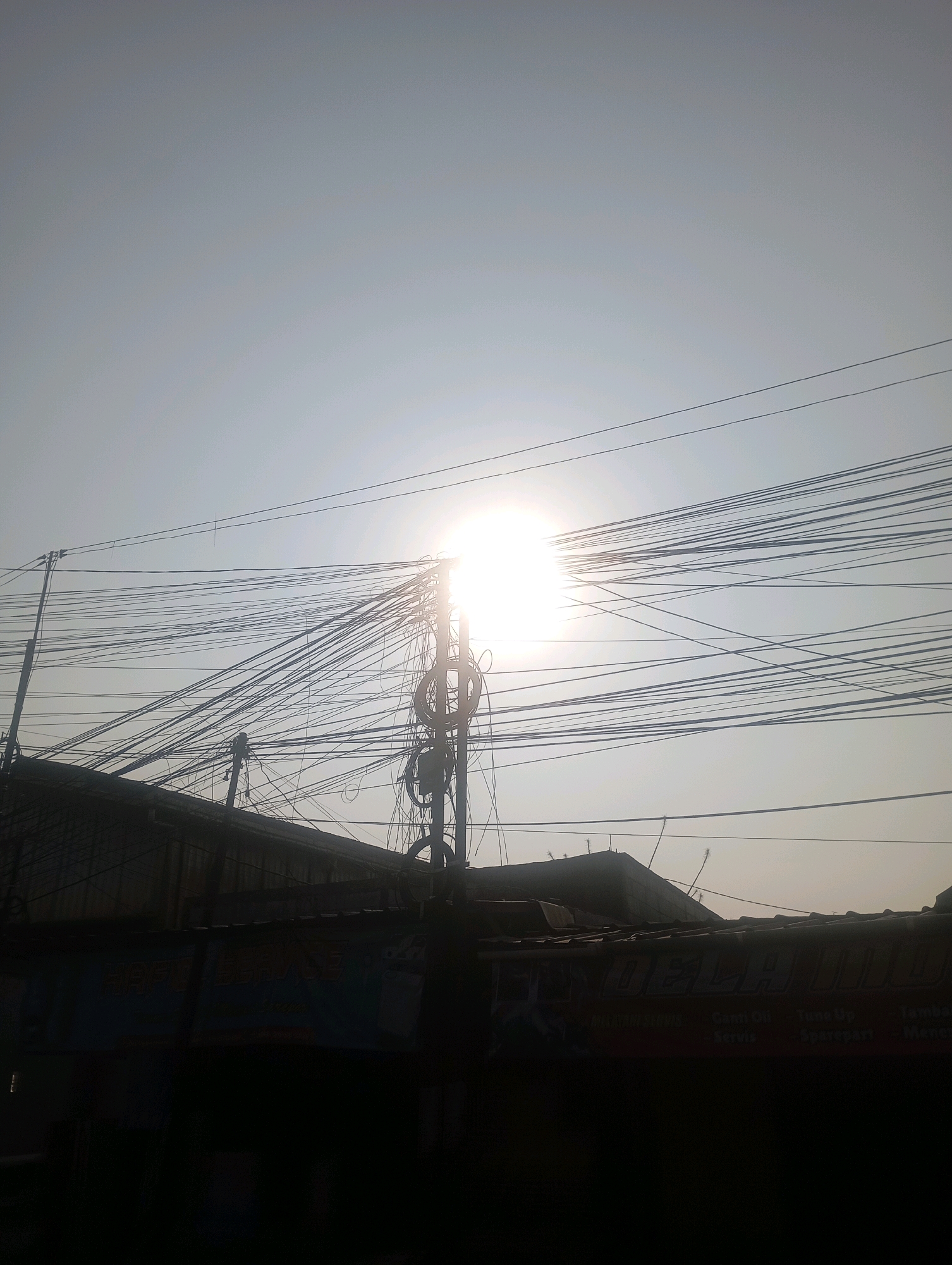 The image captures a bright, hazy sky over Babelan, Indonesia, dominated by a dense network of overhead electrical wires. The sun is positioned near the center of the frame, partially obscured by the wires and casting a strong glare. Below the wires, silhouetted against the bright sky, are the rooftops of low-rise buildings, suggesting a densely populated urban or semi-urban area. The architecture of the buildings appears to be typical of Indonesian construction, with corrugated metal or tiled roofs. One building's facade has a visible sign with text in what appears to be Indonesian, possibly advertising a business like a workshop or service center, with some words decipherable like "Bengkel" (workshop) and "Servis" (service). The overall impression is one of a bustling street scene in Babelan, characterized by functional but somewhat untidy infrastructure.