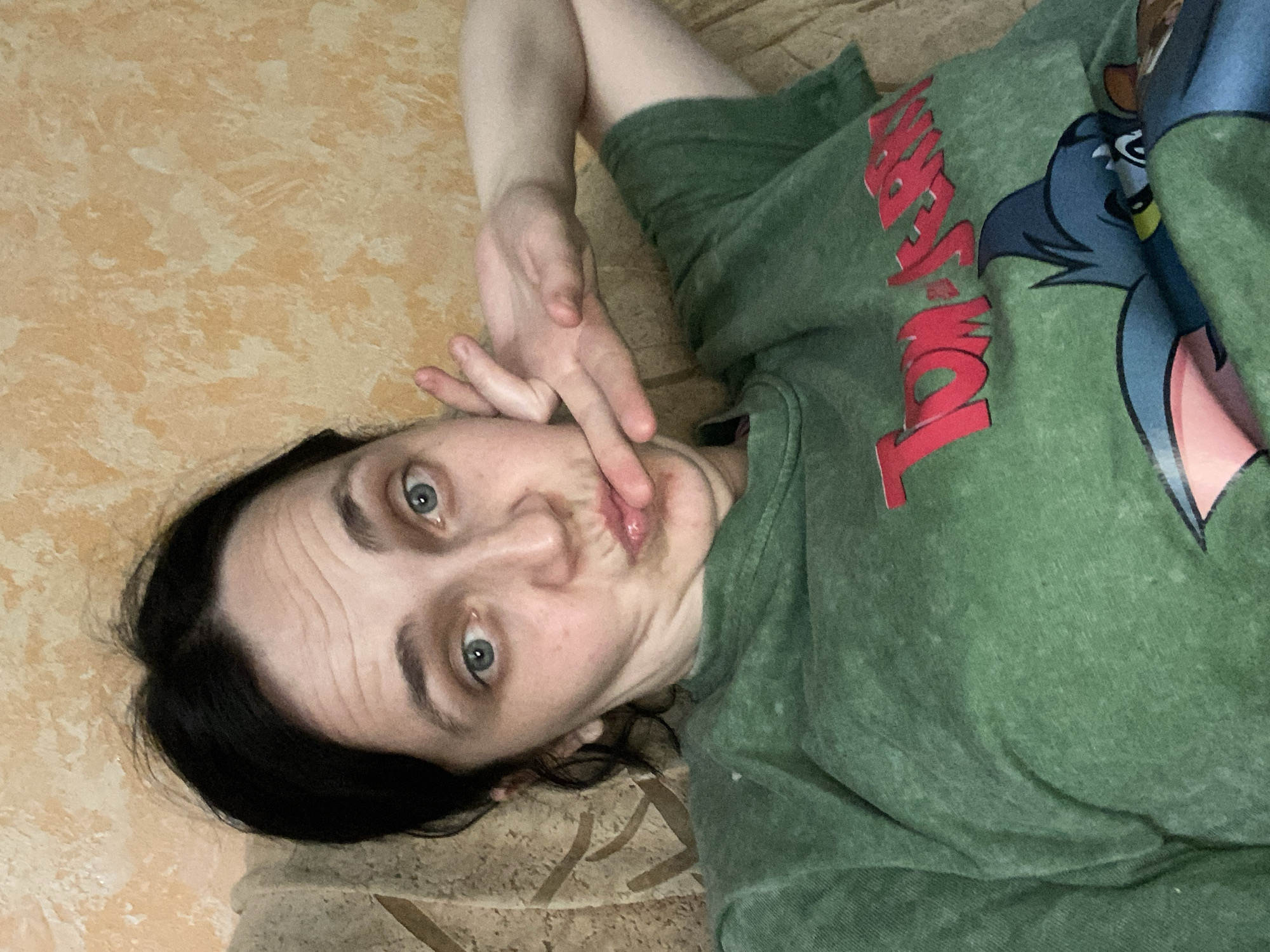 A selfie is taken of a woman with dark hair and blue eyes, who is wearing a green t-shirt with a Tom and Jerry design. She is lying down on a couch with a textured, beige-colored wall in the background. The woman is making a funny face, puckering her lips and holding up two fingers to her cheek. Her right arm is bent and resting behind her head, and her left hand is touching her face. The lighting is dim, suggesting the photo was taken indoors.