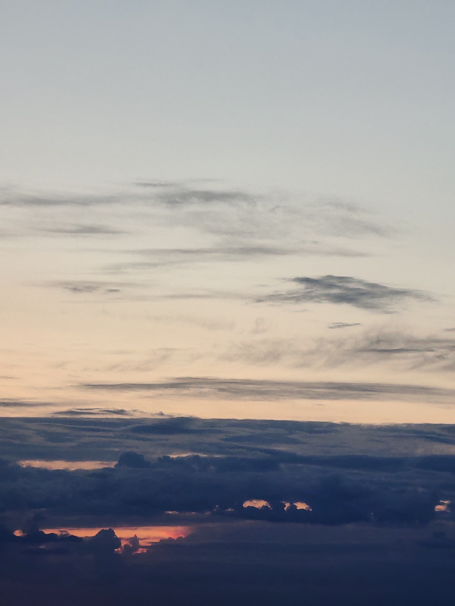 This media file captures a serene sky scene, likely at either sunrise or sunset, characterized by distinct layers of clouds and a gradient of atmospheric colors.
The upper portion of the image displays a clear, pale blue-grey sky, gradually transitioning downwards into a soft, light beige or pale orange hue. Scattered across this upper half are thin, wispy, horizontally-streaked clouds, mostly light grey in color, with some areas appearing slightly darker. These clouds are diffuse and stretched, suggesting higher altitude or atmospheric conditions that spread them out.
Below this, a prominent horizontal band of denser, darker grey clouds extends across the middle of the frame. Beneath this band, the clouds become significantly thicker and more voluminous, forming a deep, dark blue-grey layer that dominates the lower half of the image. This dense cloud cover appears to obscure the actual horizon line.
Through gaps and openings within these thick, dark lower clouds, vibrant flashes of warm light are visible. These patches are bright orange, red, and golden, strongly indicating the presence of the sun either rising or setting just below the cloud layer. The contrast between the intense warm light and the dark, heavy clouds creates a dramatic visual effect.
There are no people or specific objects discernible within the image. The scene is purely atmospheric, focusing on the interplay of light and clouds. No text is visible. The image conveys a sense of peaceful vastness and the dynamic beauty of the sky, likely observed from a vantage point looking across a wide expanse, possibly over a landscape or body of water, though these are not shown. Given the context, this sky view is from Podolsk, Russia.