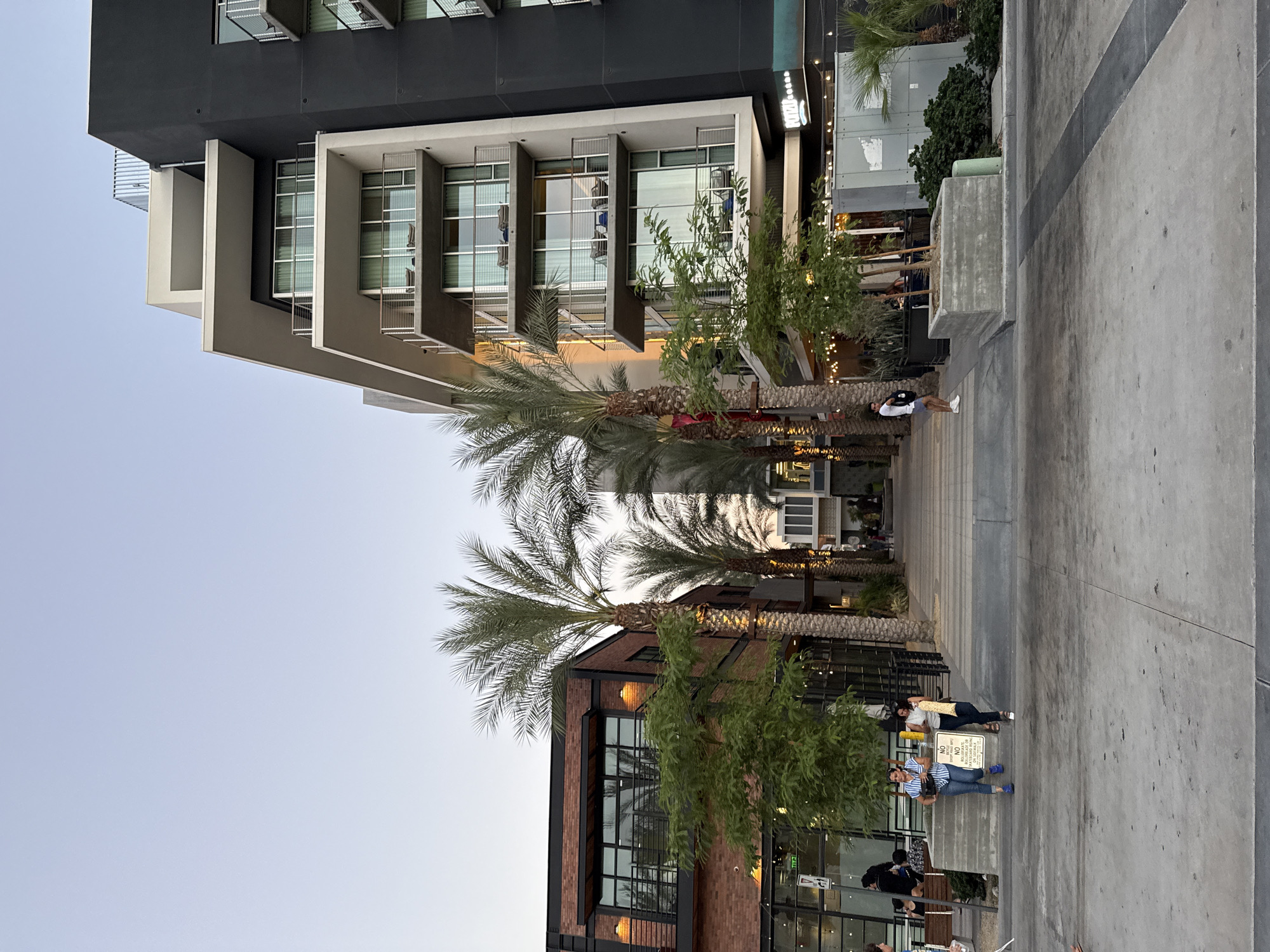 This is an eye-level, outdoor, full shot taken at dusk in Palm Springs, California, United States of America. The image captures a pedestrian walkway lined with modern buildings and palm trees. On the right is a multi-story building with a dark gray facade and balconies with glass railings. The building has a layered, geometric design. To the left is a brick building with large windows and a modern industrial aesthetic, featuring an upper-level balcony. In the center, a row of tall palm trees separates the two buildings, leading the eye down the walkway.
Several people are visible. Two women walk towards the viewer in the center foreground, one carrying a light-colored bag. A man in a white t-shirt and blue shorts stands on the left side of the walkway, facing away from the viewer. Further down the walkway, other individuals are casually strolling or seated. Outdoor lighting, including string lights over an awning, is visible, suggesting the area is a commercial or entertainment district. The sky is a pale blue, indicating the end of daylight. The ground is paved with concrete and stone tiles. A sign that reads "NO METERS VEHICLE IMPORTS" is visible near the two women.