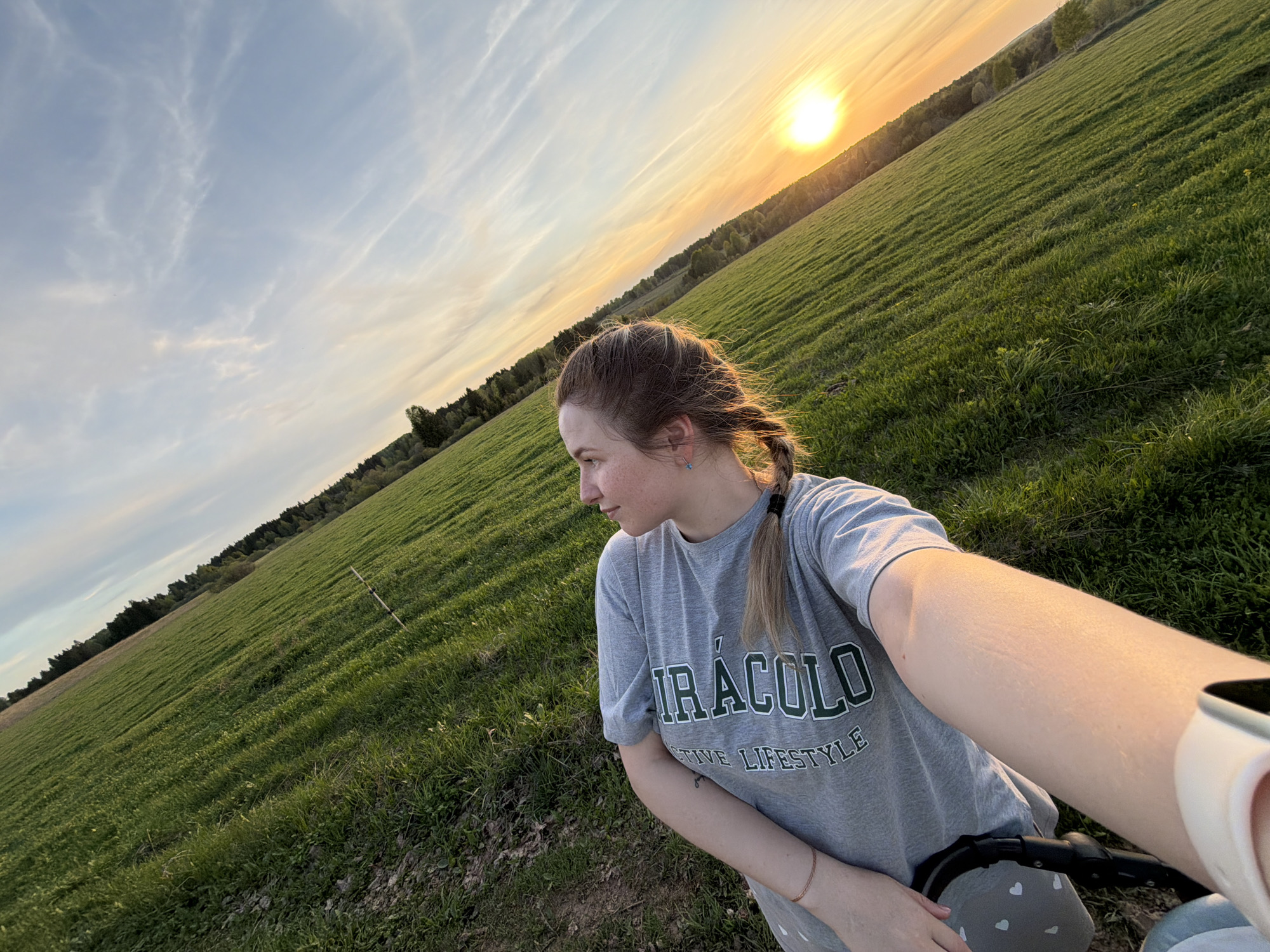The image shows a woman in an open field at sunset, possibly in the area of Брюхово, Russia. She is wearing a grey t-shirt with a logo. Her hair is styled in pigtails. She is taking a selfie with her arm extended, and she is positioned towards the left side of the frame. The background features a large green field that stretches towards a line of trees in the distance. The sun is setting, casting a warm glow over the landscape. The sky is partly cloudy with hues of blue, orange, and yellow. A small portion of a stroller is visible at the bottom of the frame.